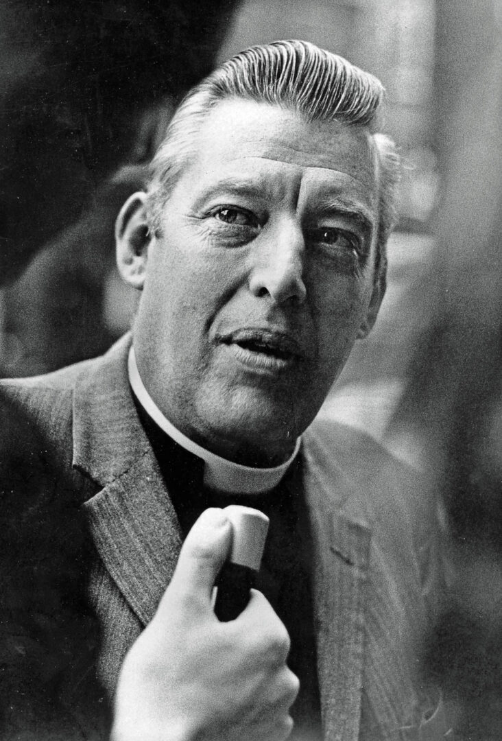 Black and white portrait of a man dressed in a suit with a clerical collar, holding a microphone and looking intently at the camera.