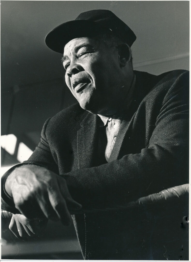 A black and white portrait of a man wearing a hat, leaning on a railing with a contemplative expression.