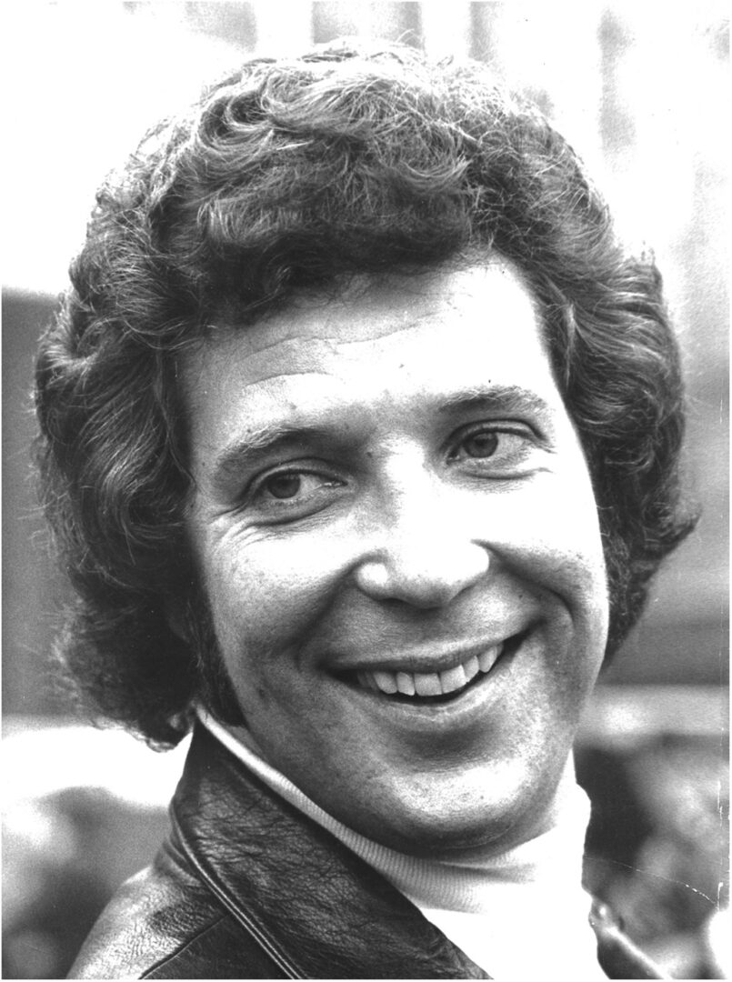 A black and white close-up portrait of a smiling man with curly hair, wearing a leather jacket and a white turtleneck.