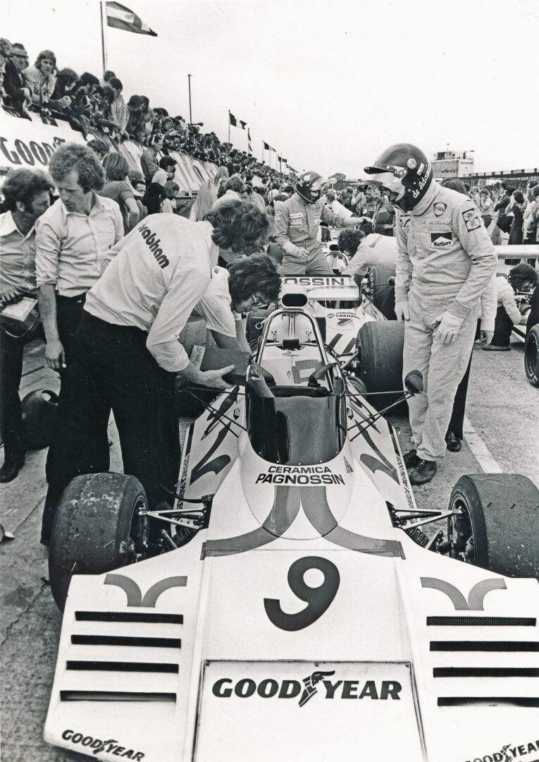A black and white photo of a racing pit stop scene, featuring a race car with the number 9 and Goodyear branding. Team members are seen working on the car, while a driver in a helmet stands nearby. Spectators are visible in the background, watching the action.
