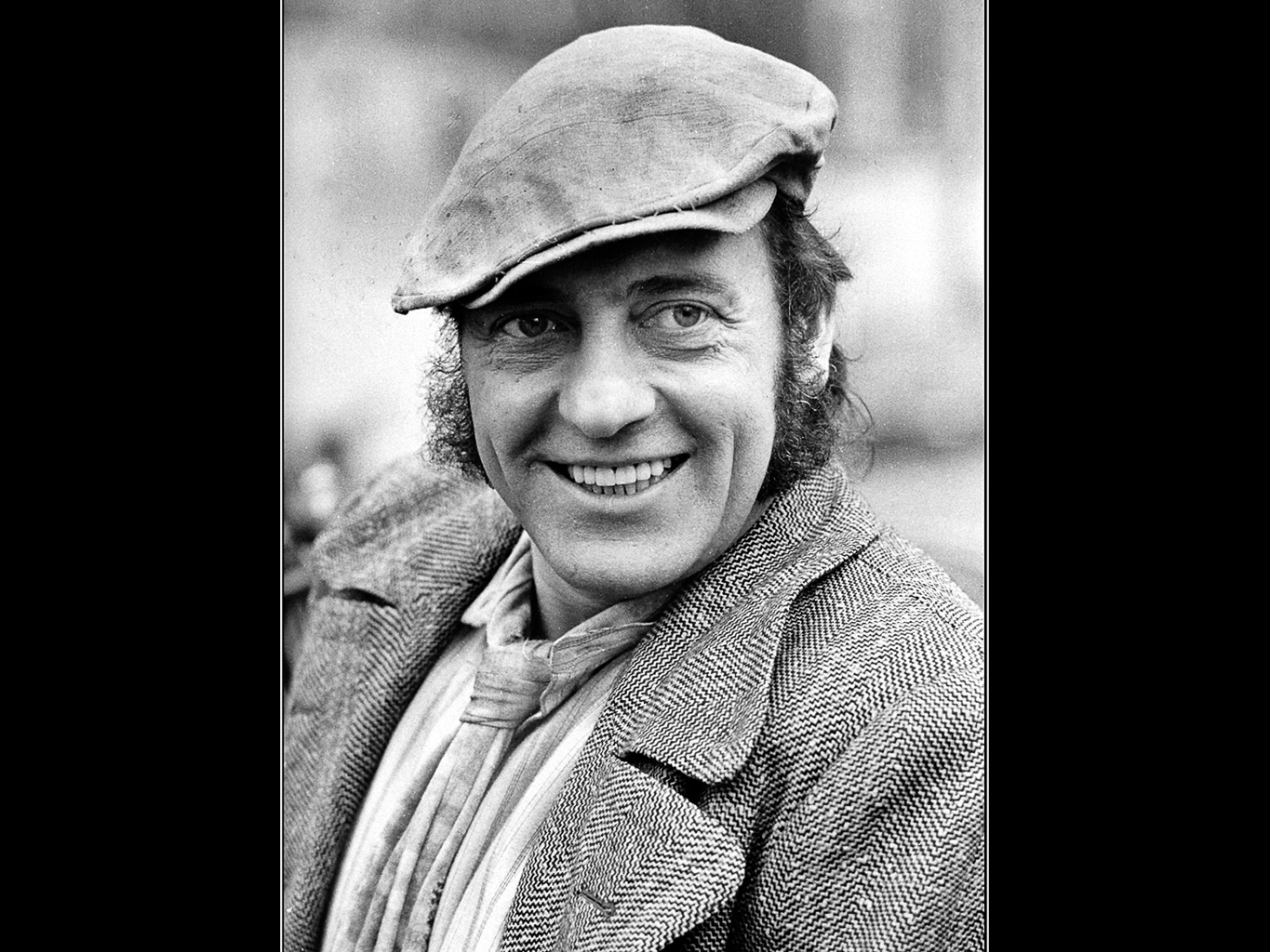 A smiling man wearing a flat cap and a patterned coat, with curly hair visible, captured in a black and white portrait.