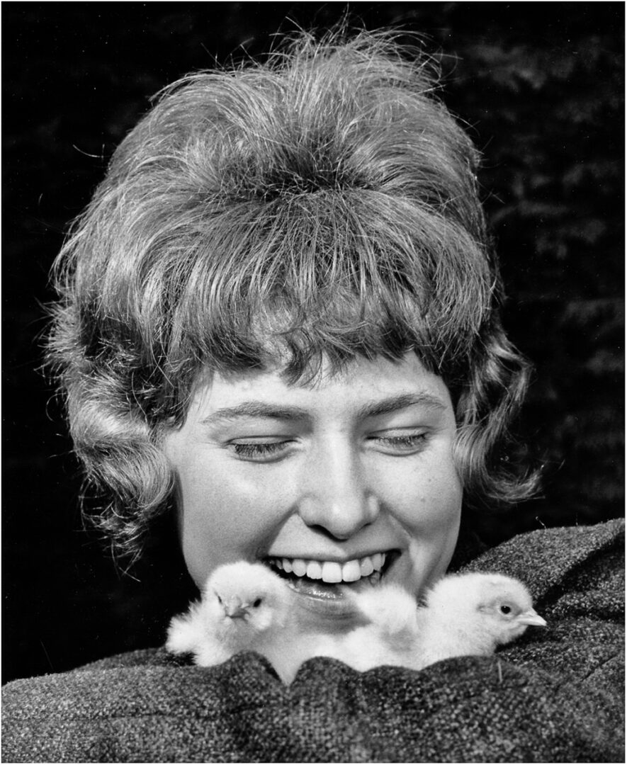 A smiling woman with curly hair holds two fluffy chicks in her lap, capturing a joyful moment.