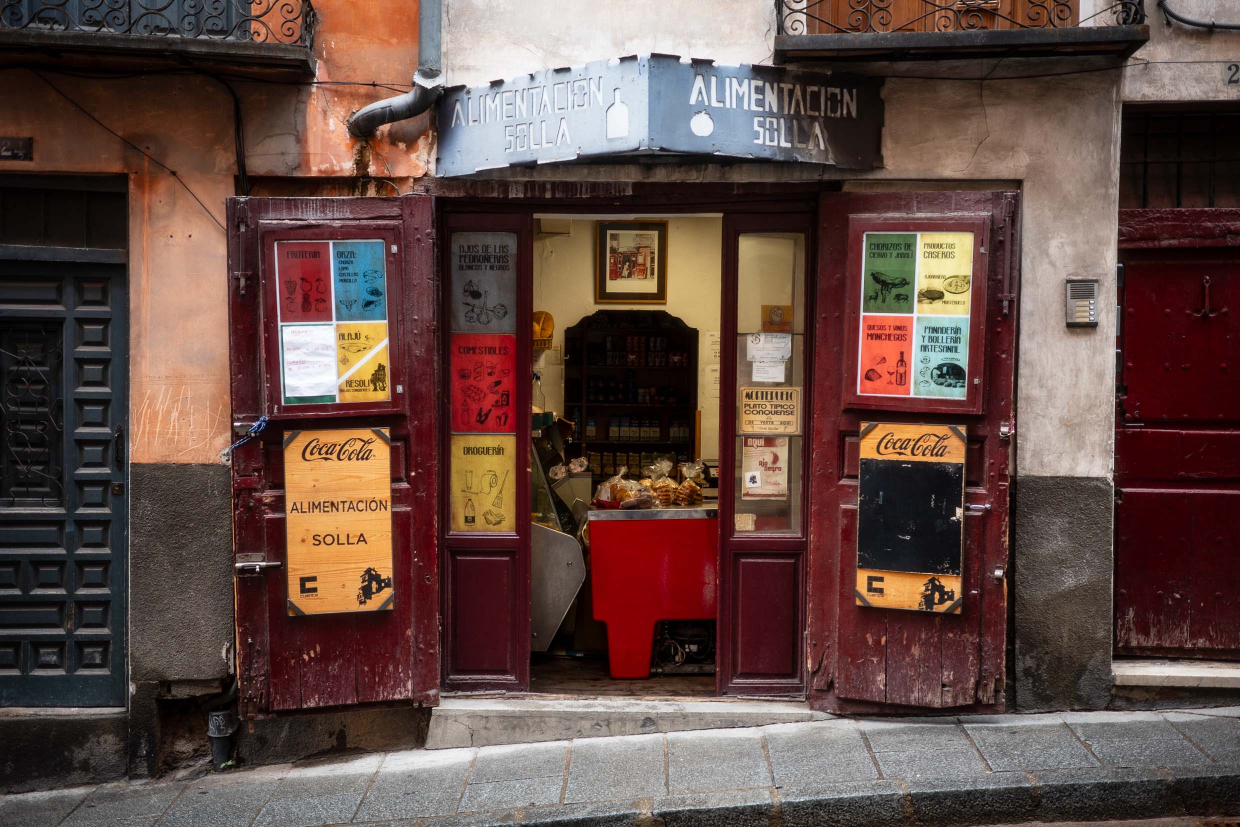 Exterior view of a small local grocery store named 'Alimentación Solla', featuring wooden double doors and colourful advertisements, including Coca-Cola signage. The interior is partially visible with shelves stocked with goods.