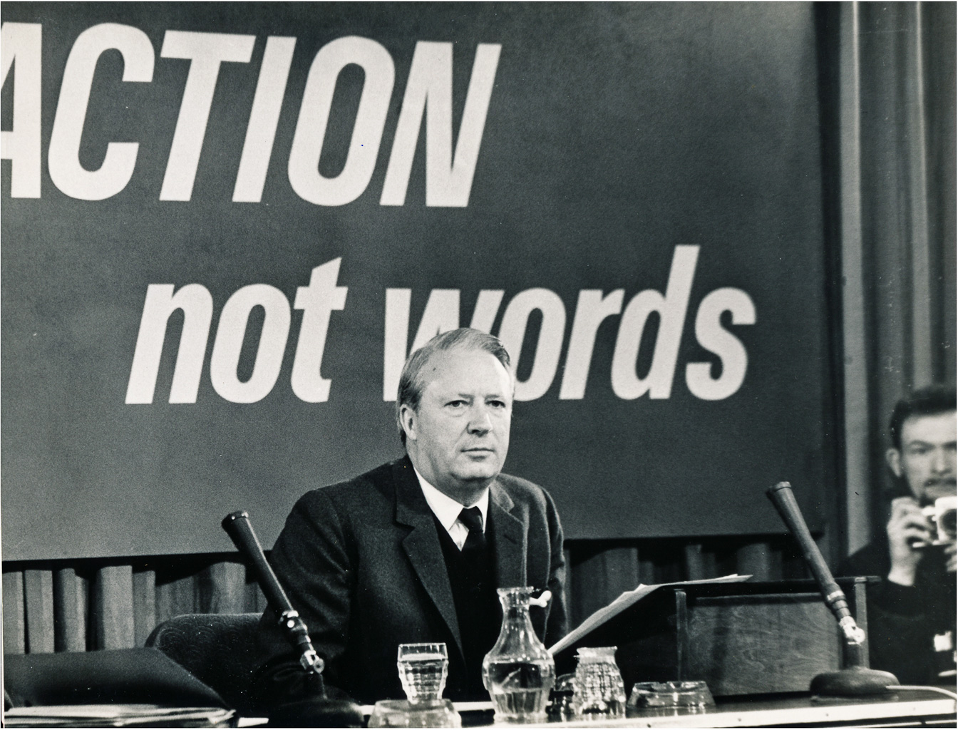 A black and white photograph of a man seated at a conference table, speaking into a microphone. Behind him, a large banner reads 'ACTION not words'. In the background, another person is visible, holding a camera.
