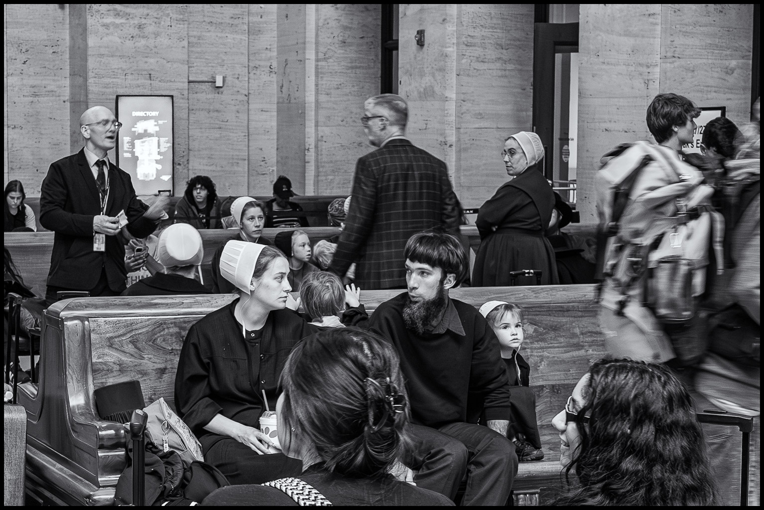 A black and white image of a busy public space, featuring a mix of people, including a man in a suit speaking, a woman in traditional dress, and children, seated and interacting, while others move through the scene.