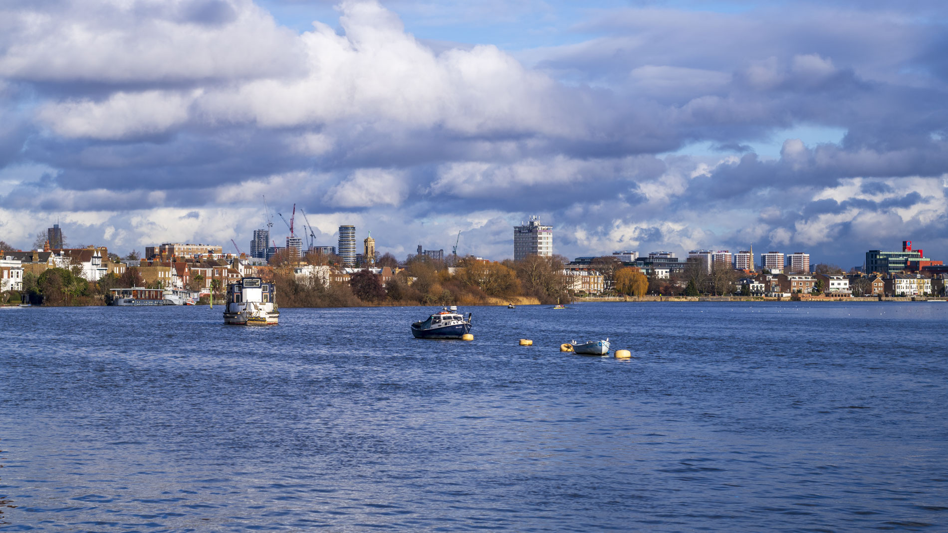 A scenic view of a river with several boats, surrounded by city buildings and blue skies with clouds.