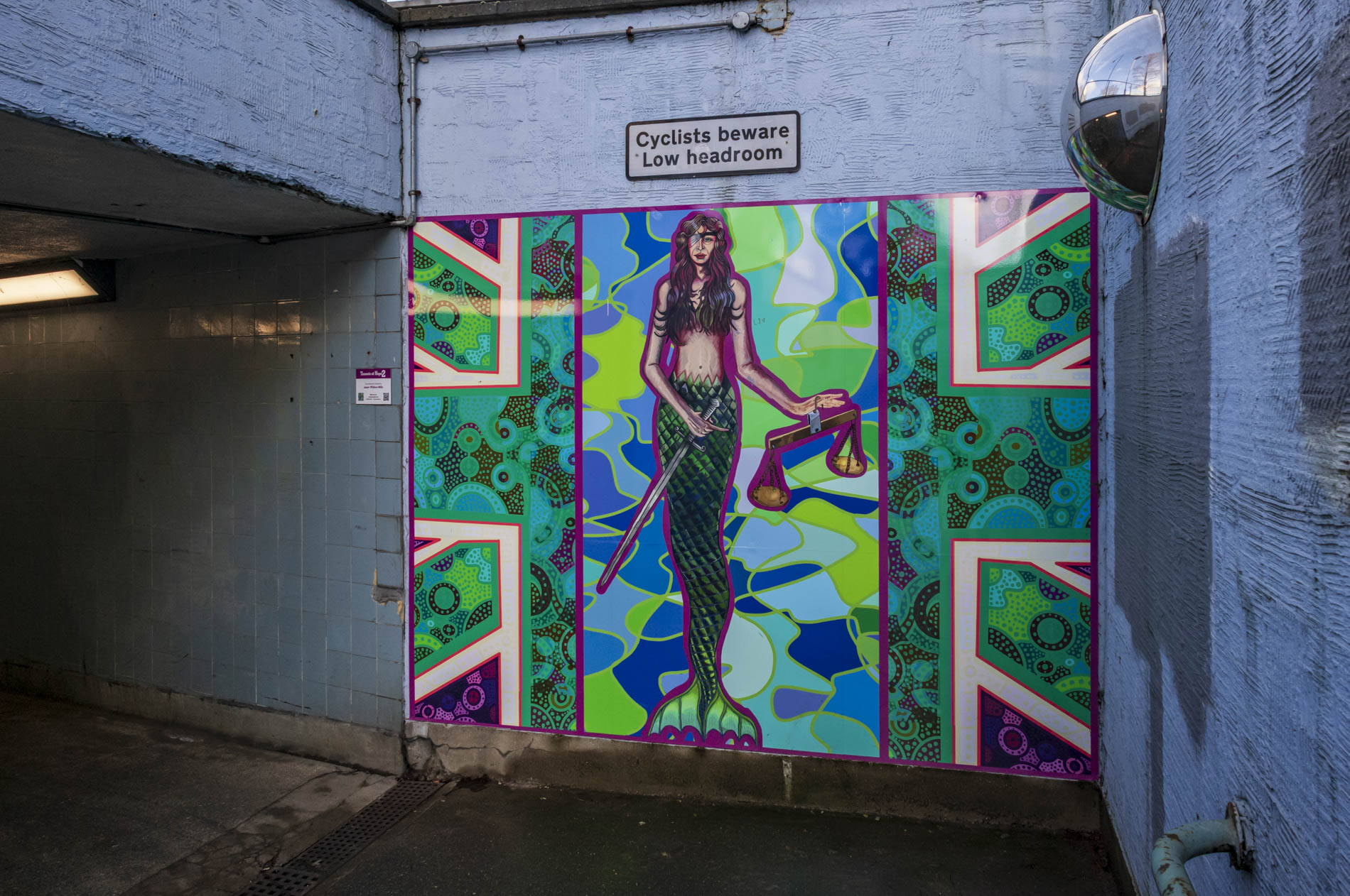 A colourful mural featuring a mermaid holding scales, surrounded by vibrant geometric patterns and colours. Above the mural, a warning sign reads 'Cyclists beware Low headroom'.
