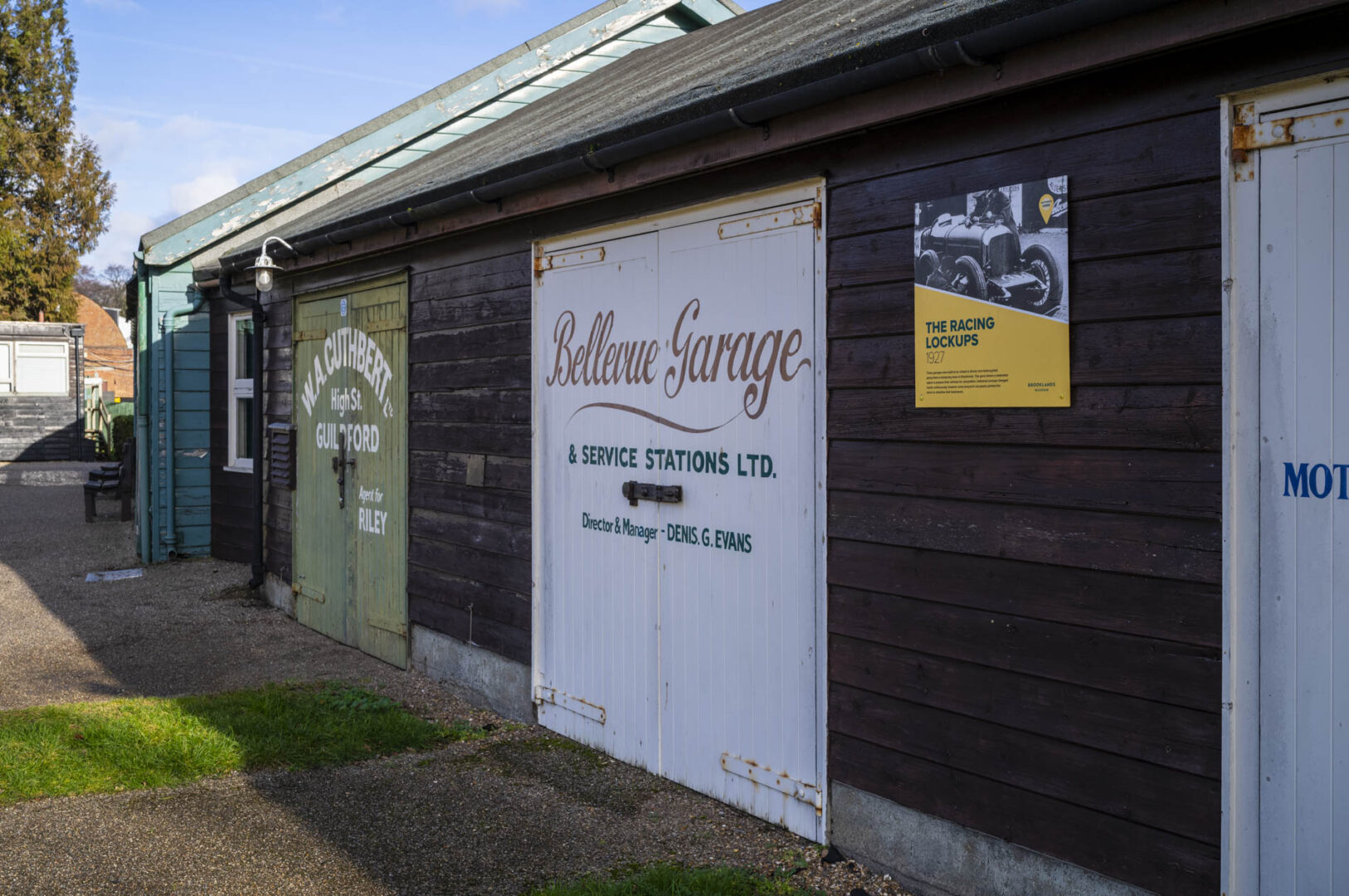 Exterior view of Bellevue Garage and service station with wooden doors and signage, featuring a poster about 'The Racing Lockups' on the wall.