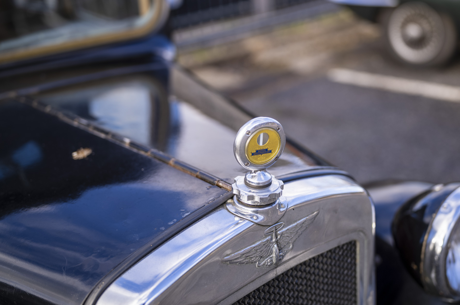 Close-up of a vintage car's bonnet ornament featuring a yellow and silver thermometer.