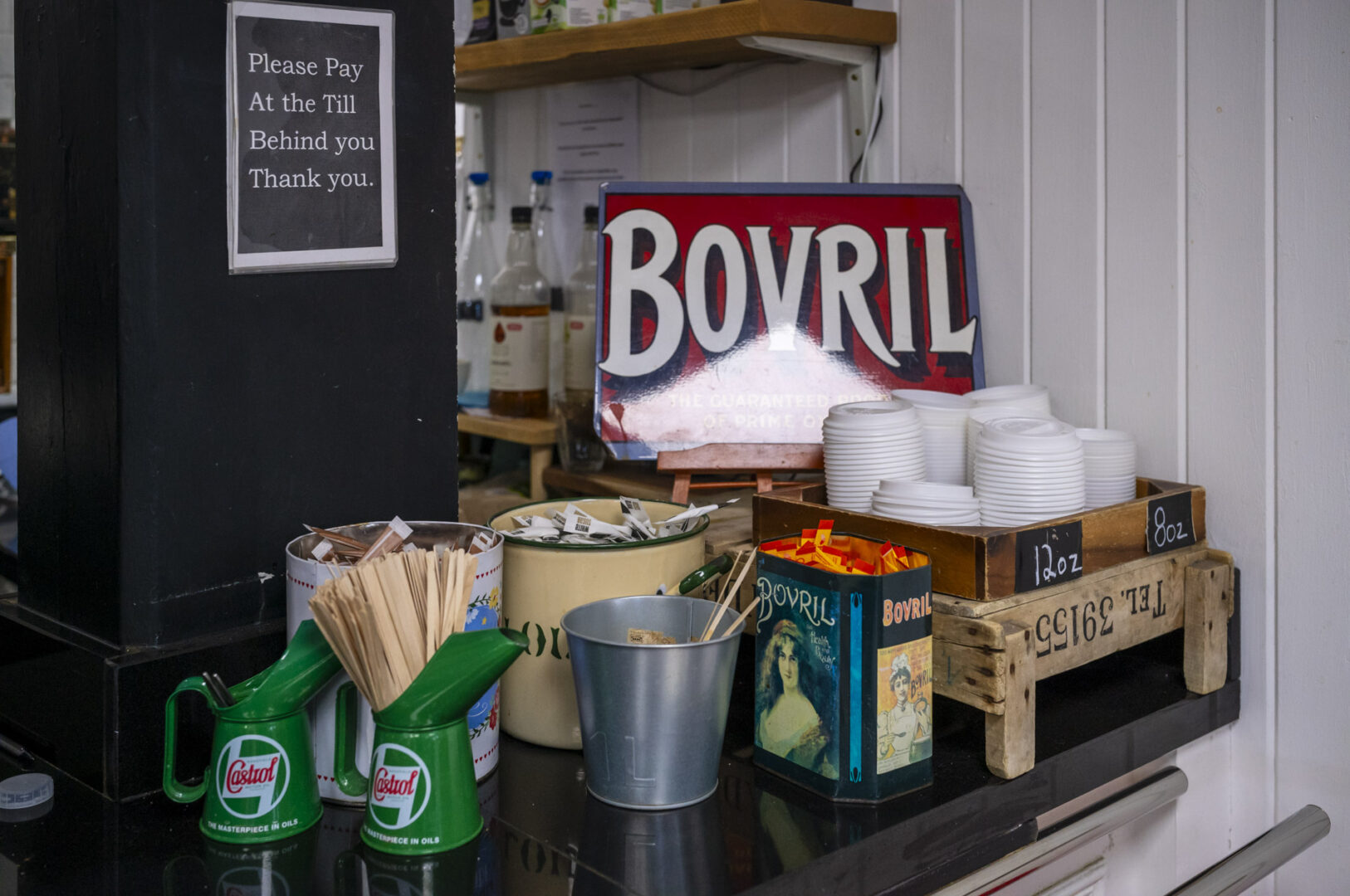 A cafe counter featuring a 'Bovril' sign, green condiment dispensers, wooden crates, and disposable cups. A sign instructs customers to pay at the till behind.