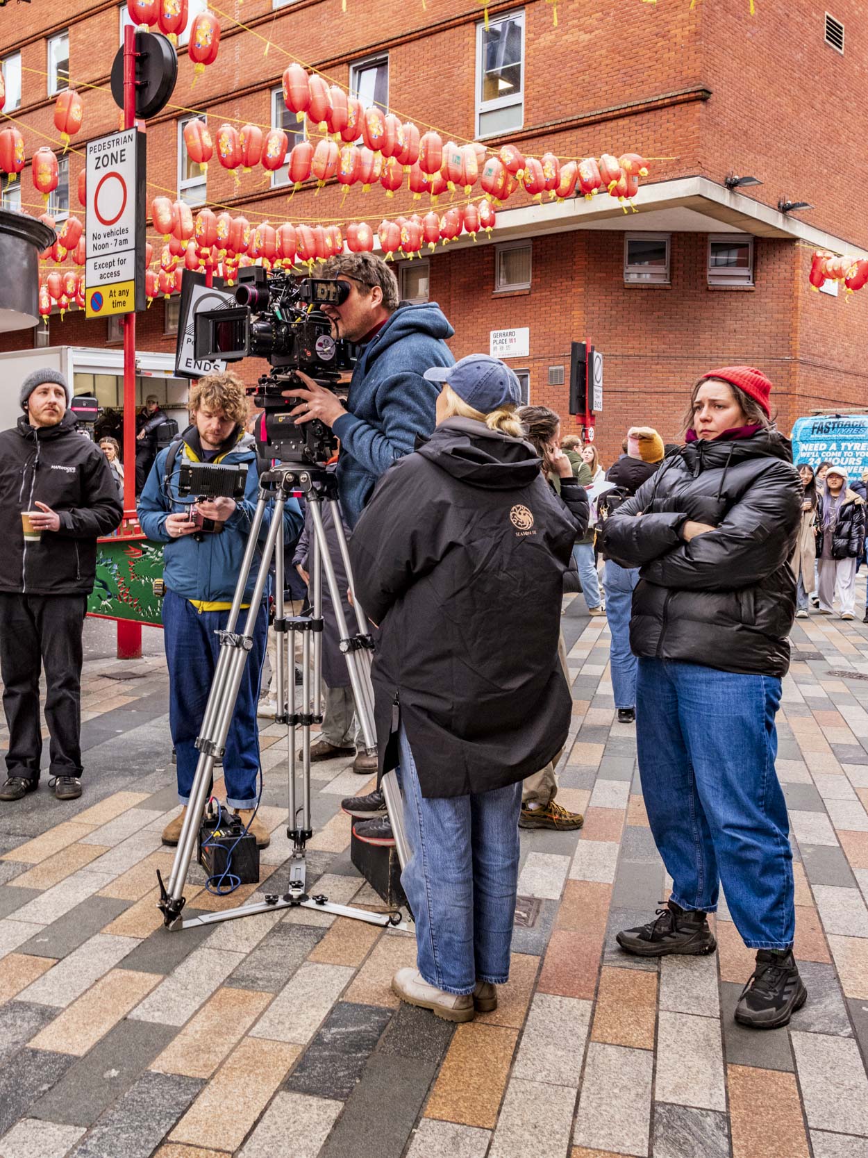 A film crew is set up in a pedestrian area with red lanterns overhead. A cameraman operates a camera on a tripod while crew members observe, holding drinks and wearing casual clothing.