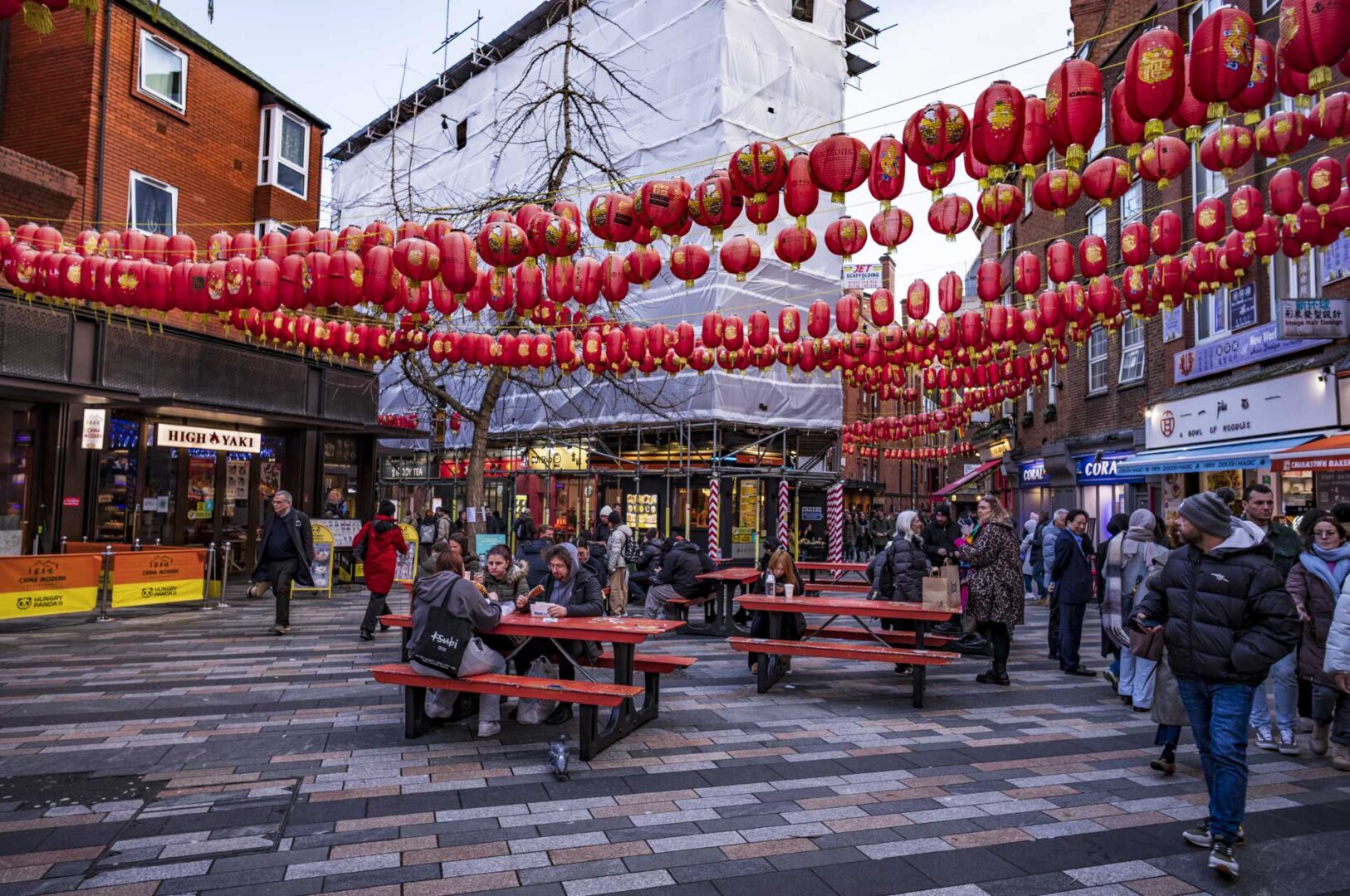 A bustling street with red Chinese lanterns hanging overhead, people walking and sitting at red picnic tables, surrounded by shops and a construction site in the background.