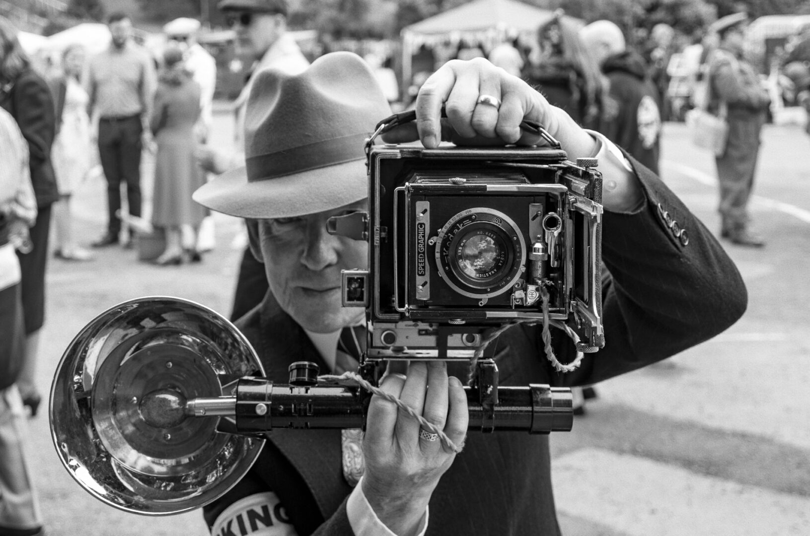 A man in a vintage outfit wearing a hat, holding an old-fashioned camera with a large lens and bellows while pointing it toward the viewer. The setting is outdoors with a crowd of people in the background.
