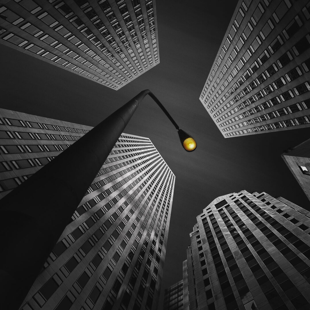 A low-angle view of tall skyscrapers reaching towards the sky, with a street lamp illuminated in yellow in the foreground against a monochrome backdrop.