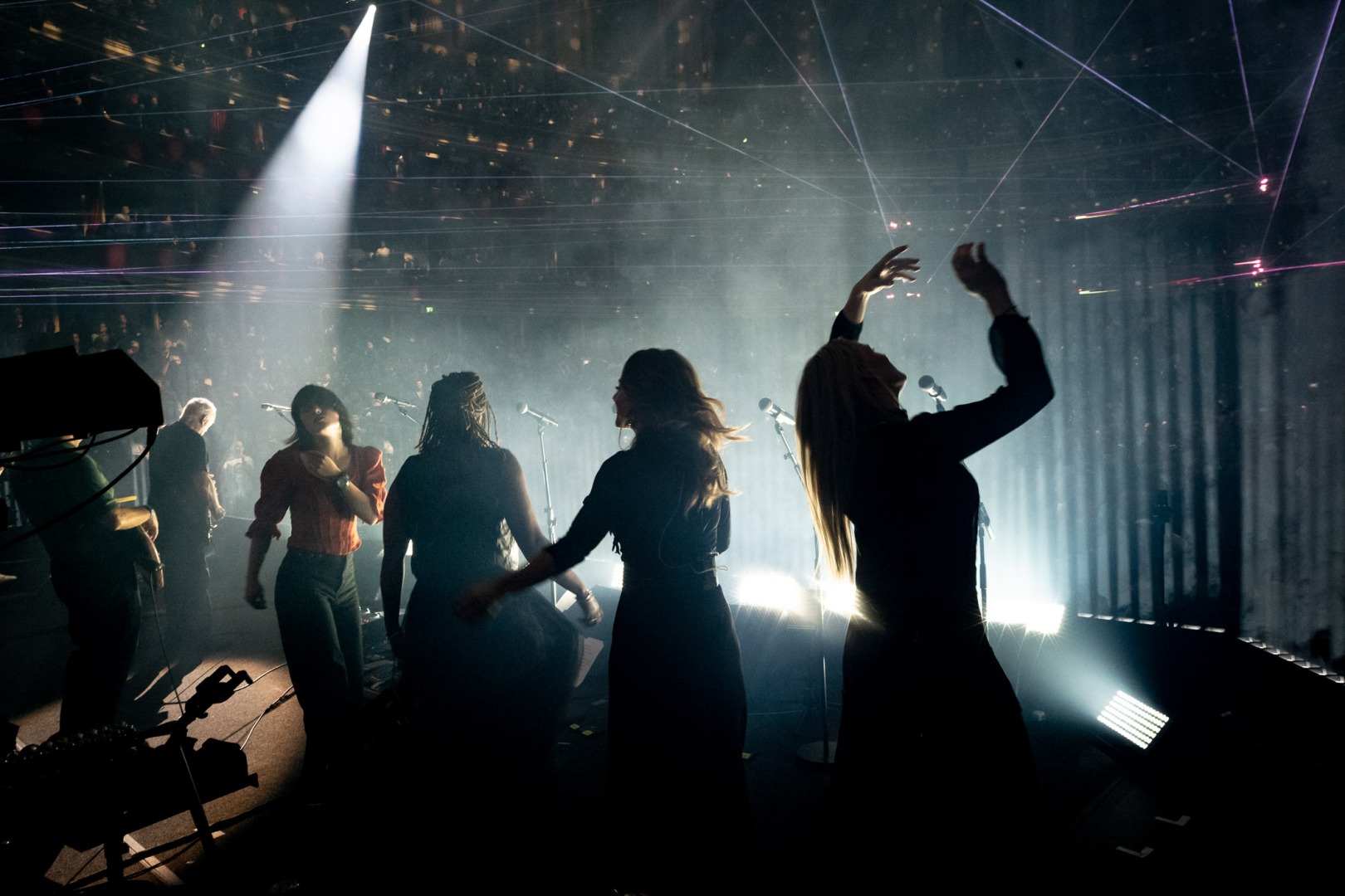 A group of performers on stage, silhouetted against dramatic lighting and laser effects, with a captivated audience in the background.