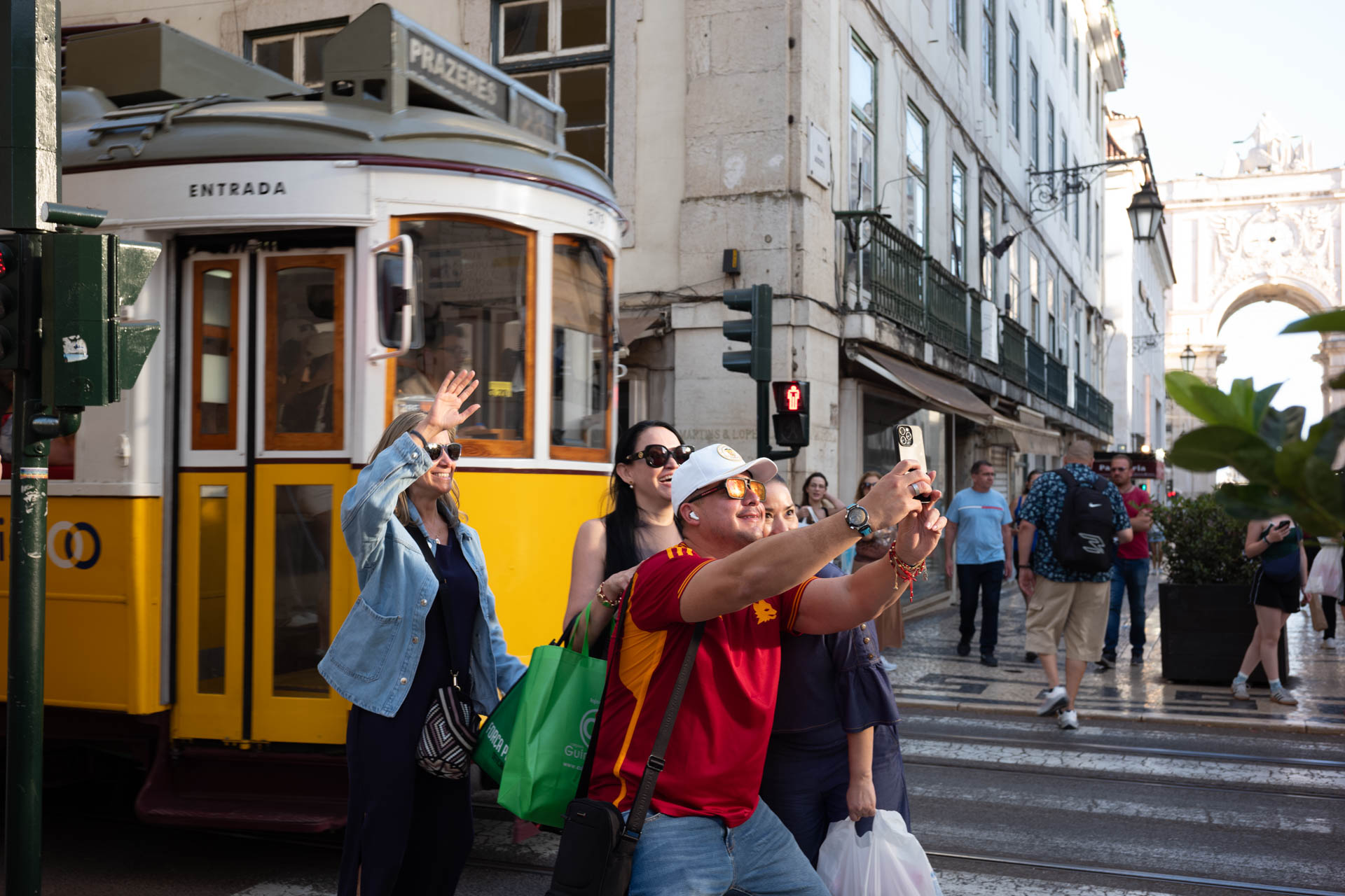 A group of people taking selfies in front of a vintage yellow tram in a busy city street, with one person waving and others smiling.