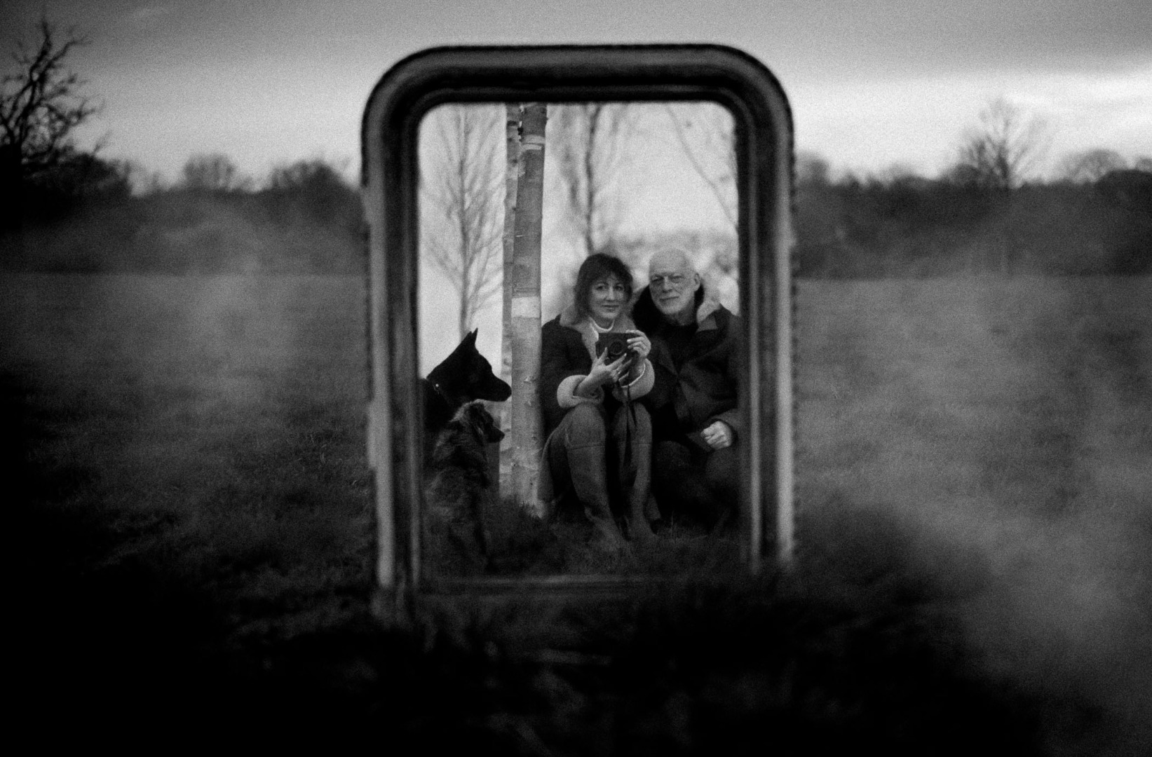A black and white photograph of a couple sitting together in a field, reflected in an antique mirror. They are holding a camera, with a dog sitting beside them.