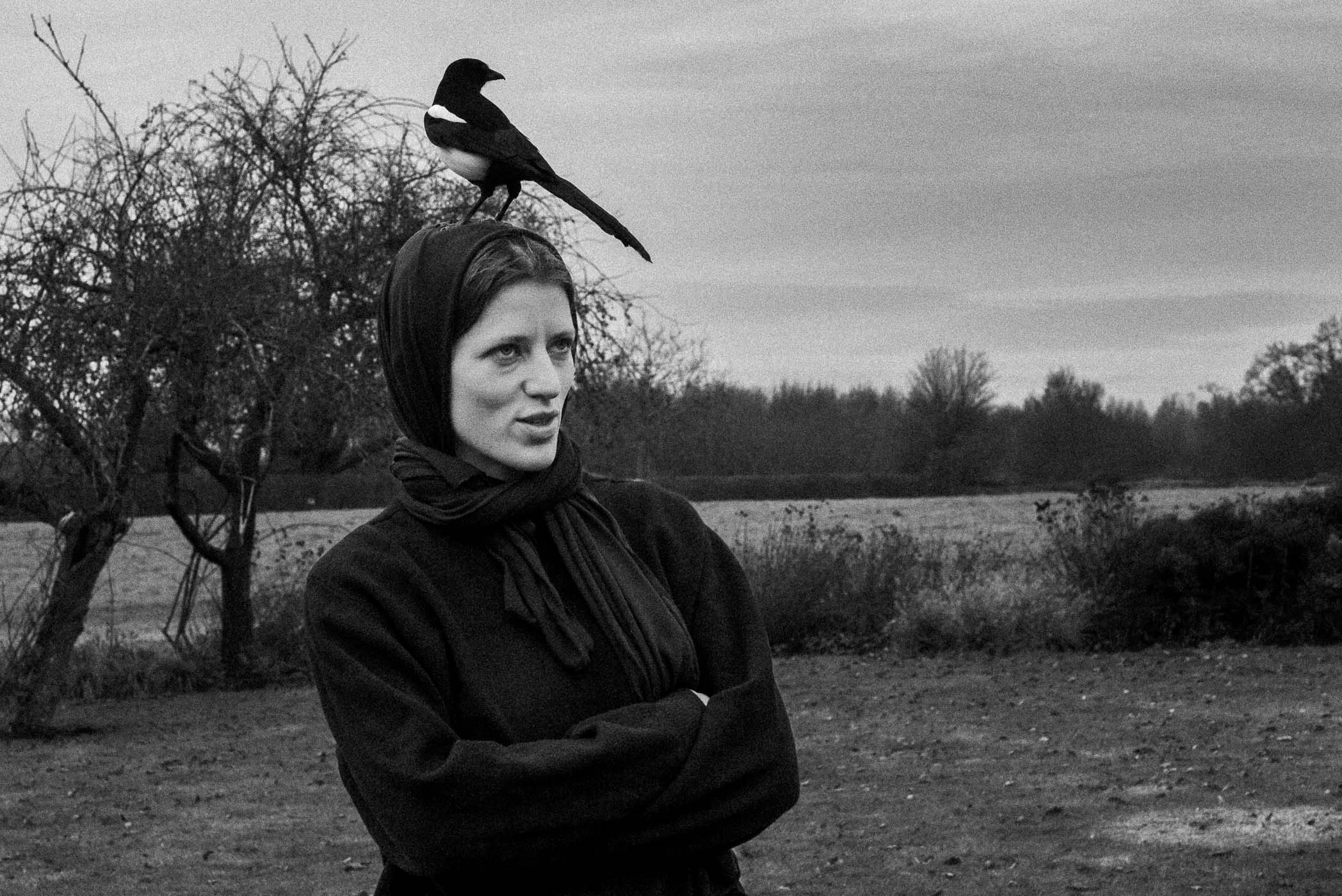 A woman in a black coat and scarf stands outdoors with her arms crossed, a magpie perched on her head, against a backdrop of trees and a cloudy sky.