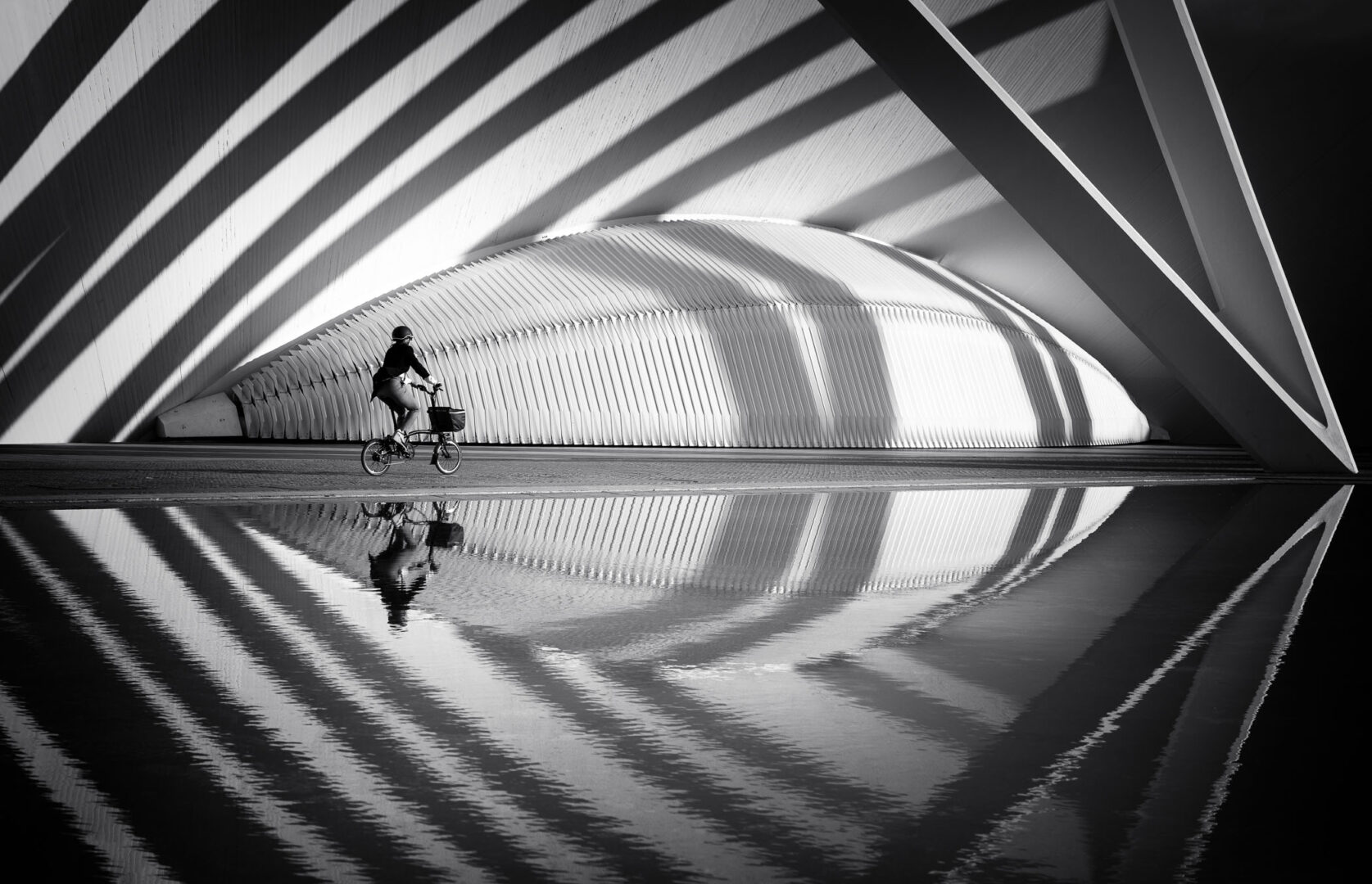 A person riding a bicycle near a striking modern architectural structure, reflected in a pool of water. The scene is in black and white, showcasing dramatic shadows and lines.