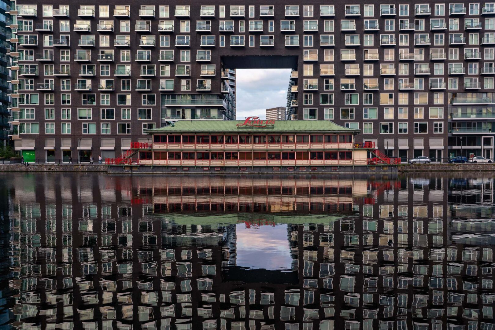 A modern building with a large open square and numerous windows reflecting in the water, with a traditional boat house in the foreground.