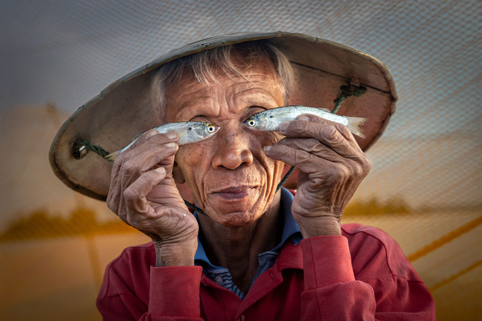 An elderly man wearing a conical hat holds a small fish in each hand in front of his face, smiling at the camera.