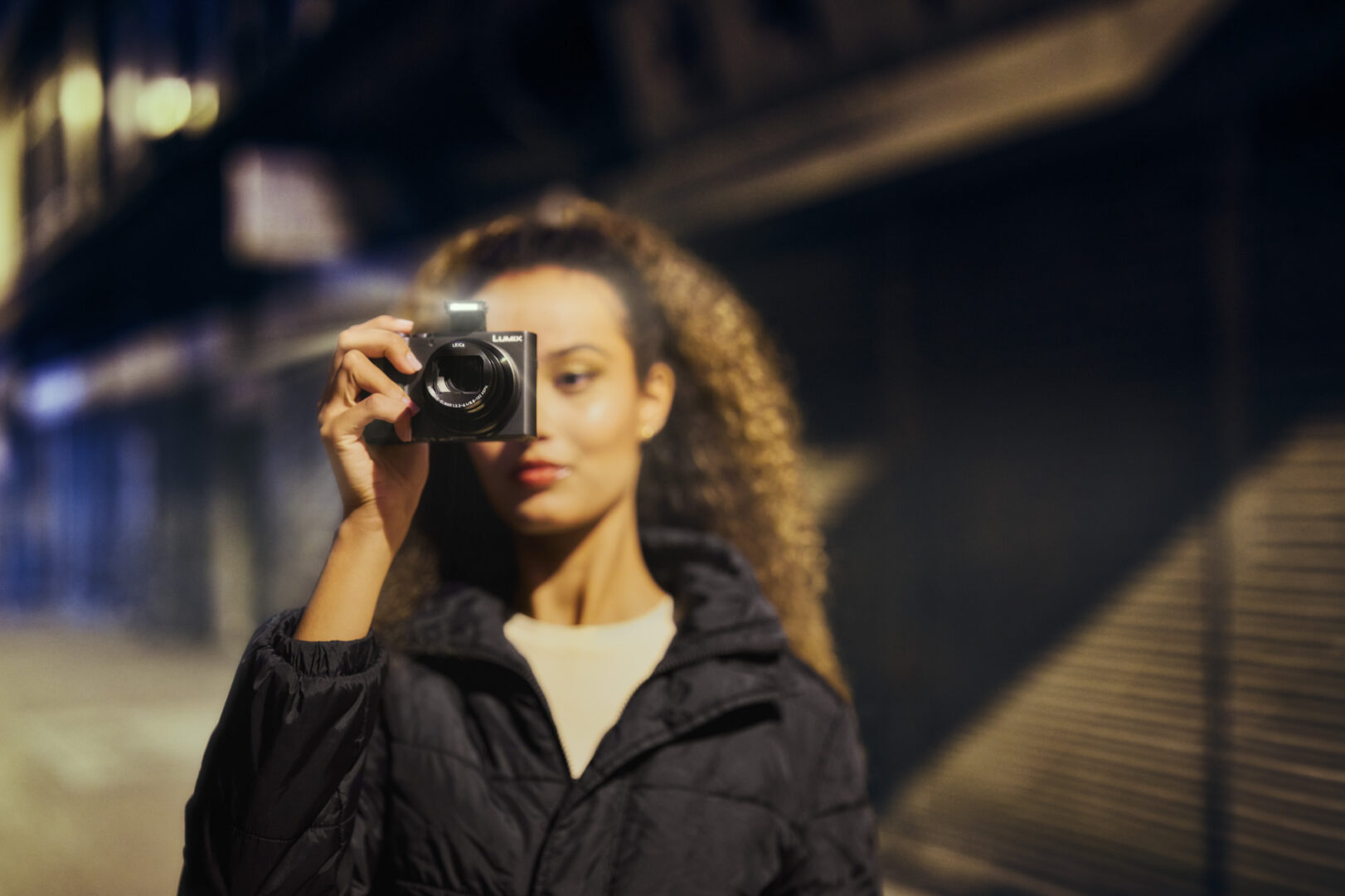 A woman holding a camera in a dimly lit urban street, wearing a black jacket with curly hair.