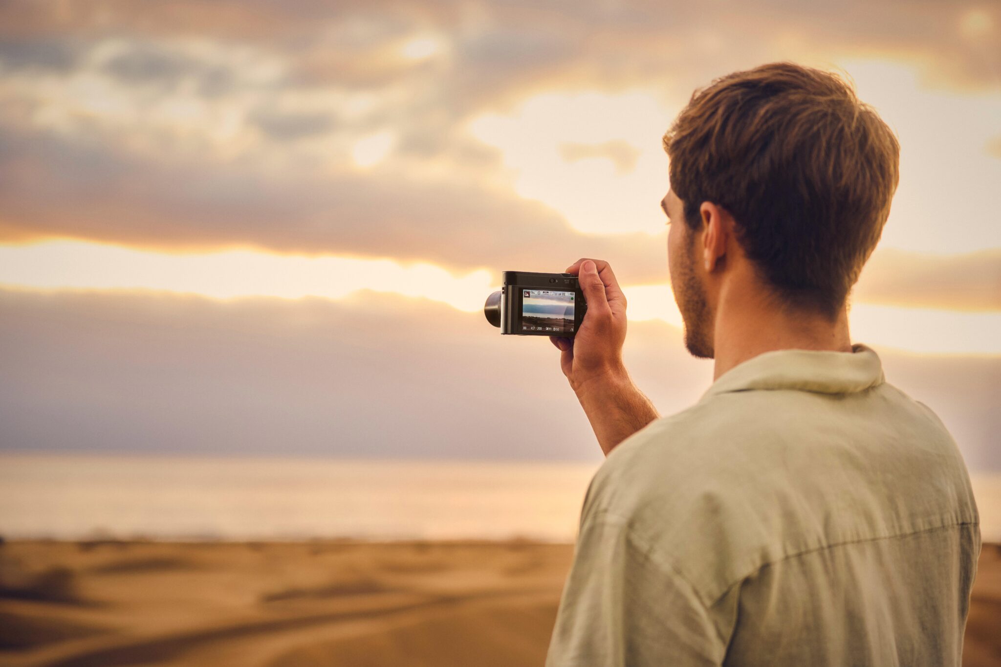 A man standing on sand dunes, holding a camcorder while capturing a scenic sunset over the ocean.
