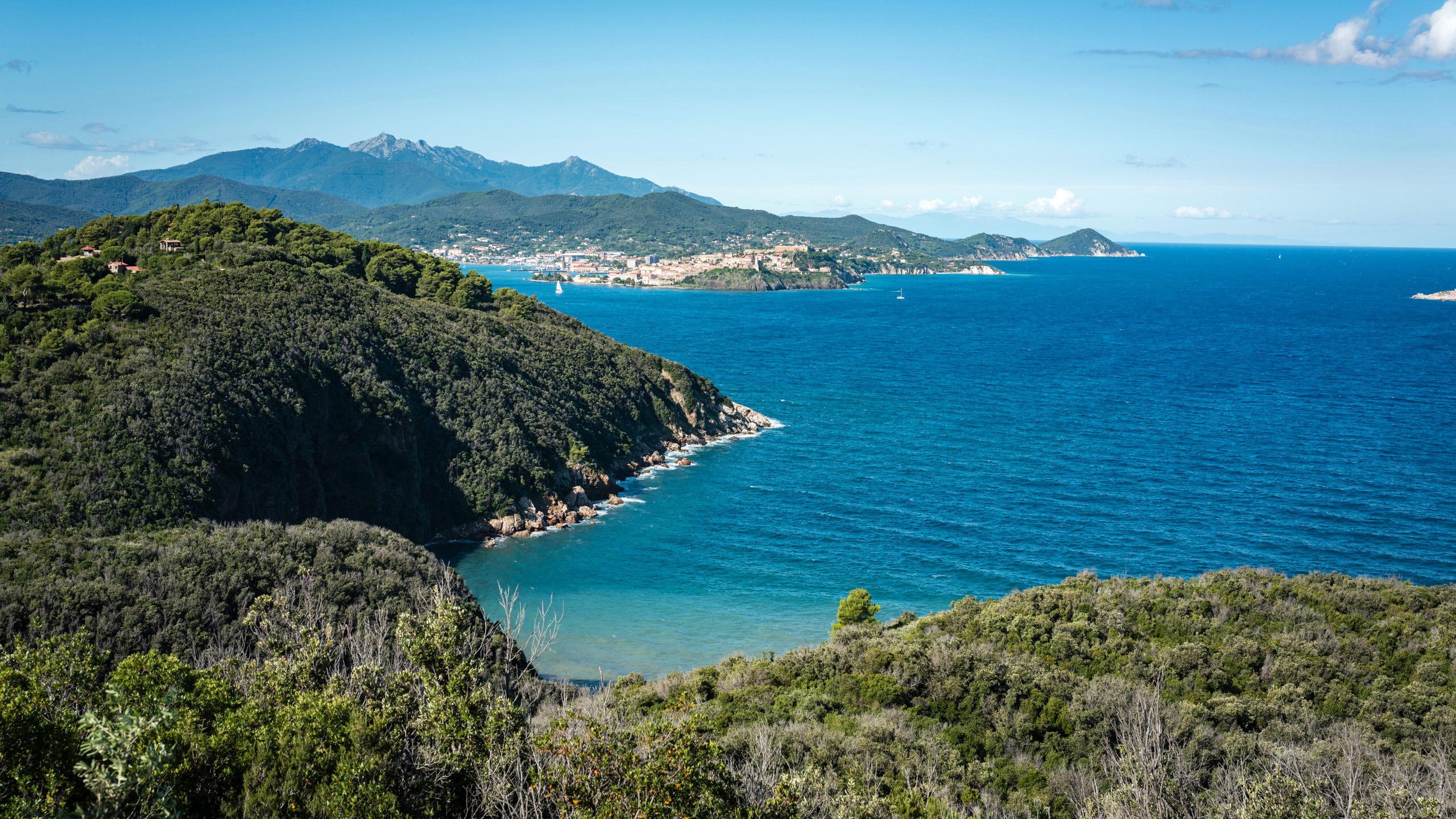 A panoramic view of a coastal landscape featuring lush green hills, a blue sea, and distant mountains under a clear sky.