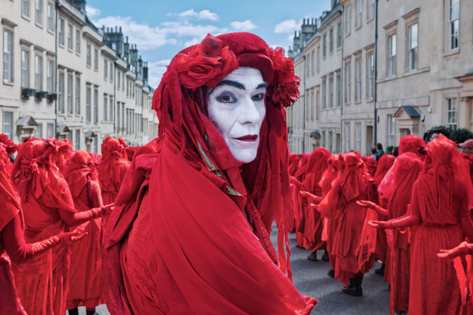 A performance artist in a striking red costume and white-painted face stands out among a crowd of similarly dressed individuals in an urban setting.