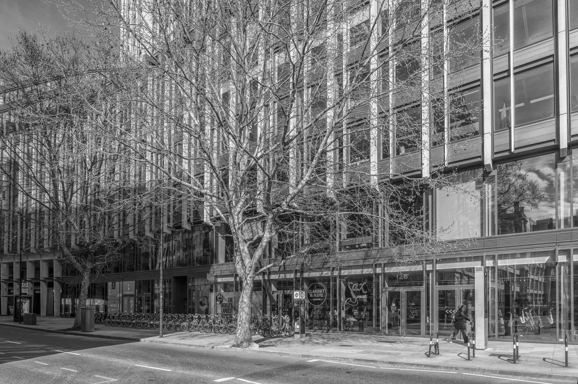Black and white image of a modern building with glass windows and a tree in the foreground, showing a person walking past a storefront.