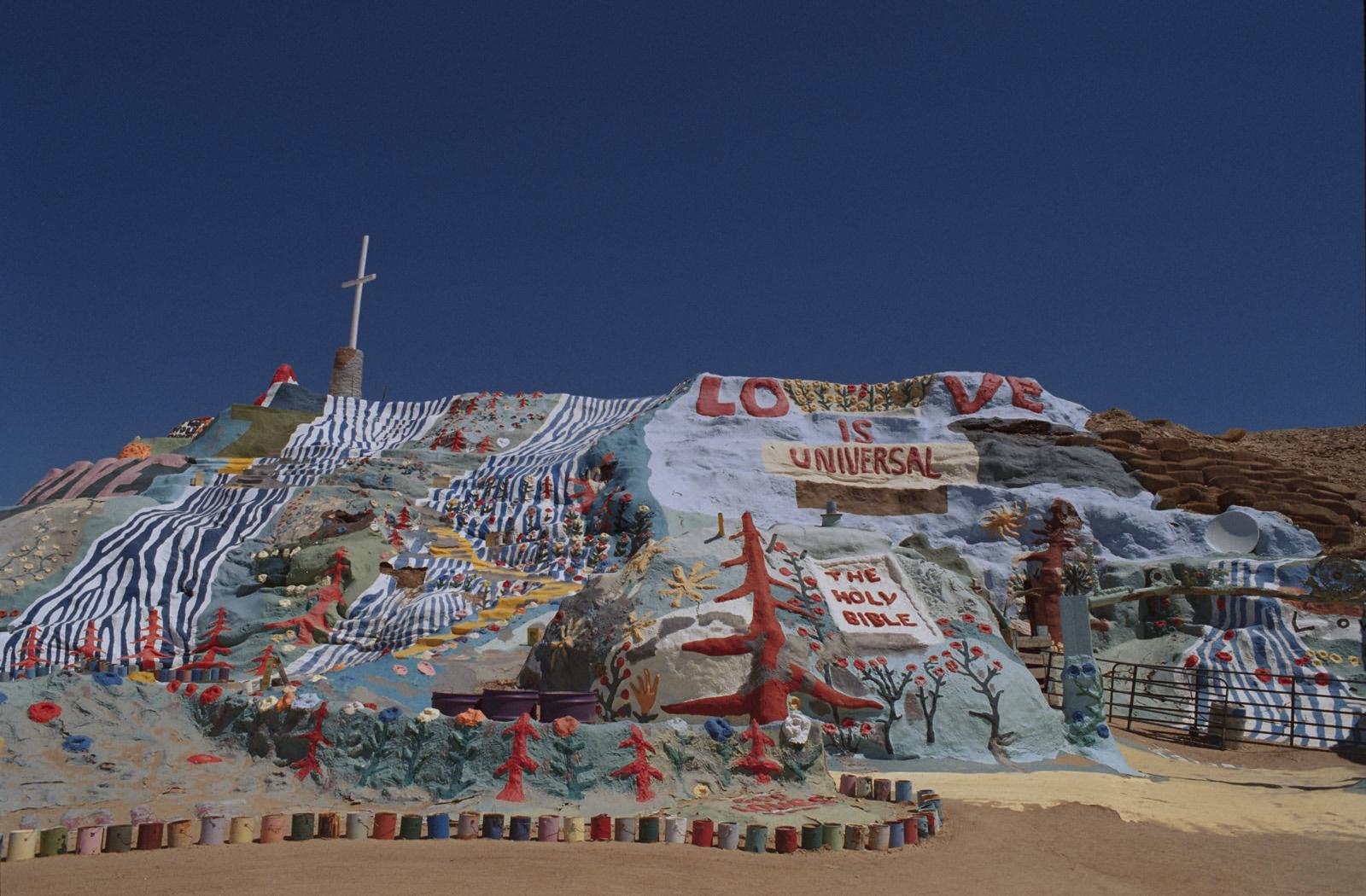 Colourful hillside mural featuring a large cross, various painted designs, and text stating 'Love is Universal' and 'The Holy Bible'.