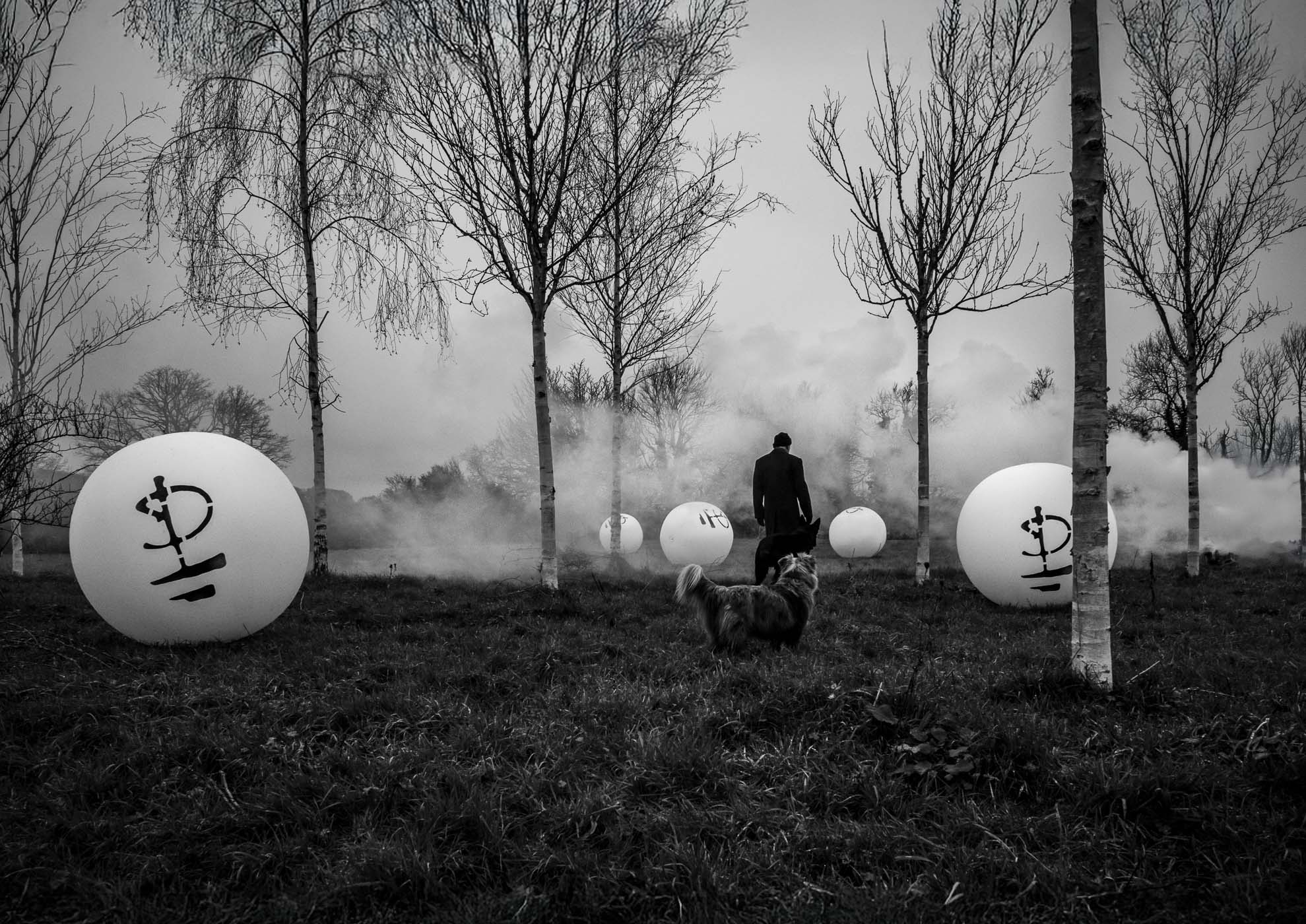 A figure walking away among large, white spherical objects with symbols, surrounded by trees and mist, creating a surreal atmosphere in black and white.