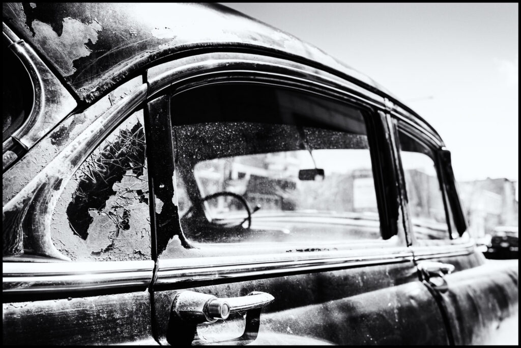 CL JC Close-up of an old, dusty car window with peeling paint and reflection of a cityscape.