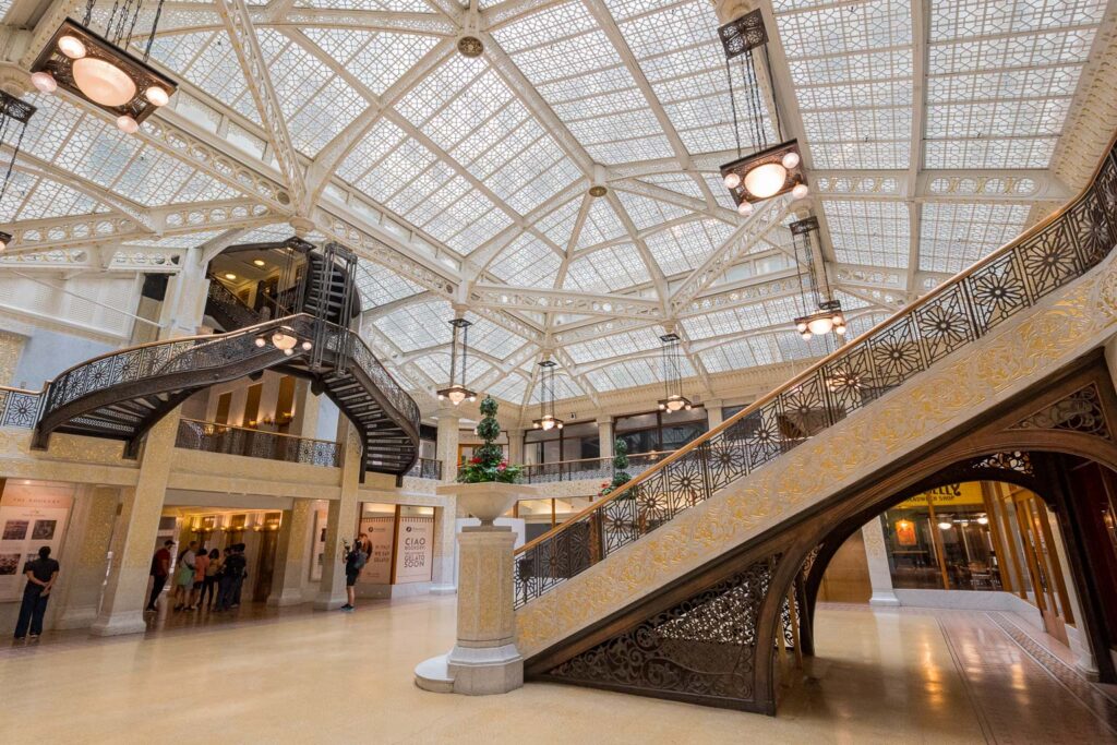 Interior view of a historic building featuring ornate staircases, a glass ceiling, and decorative lighting.