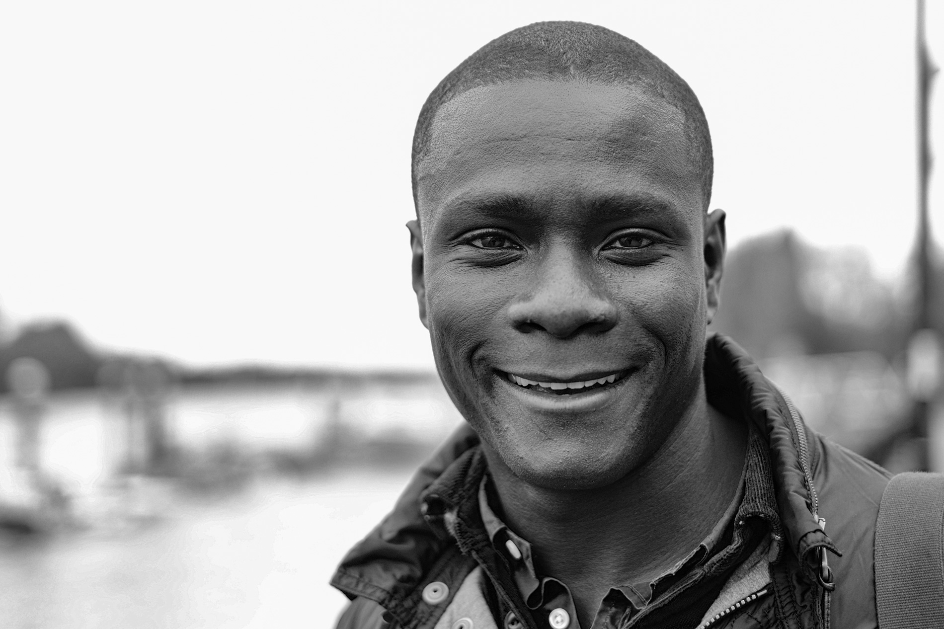 A smiling man with short hair, wearing a jacket and standing near a river, in black and white.