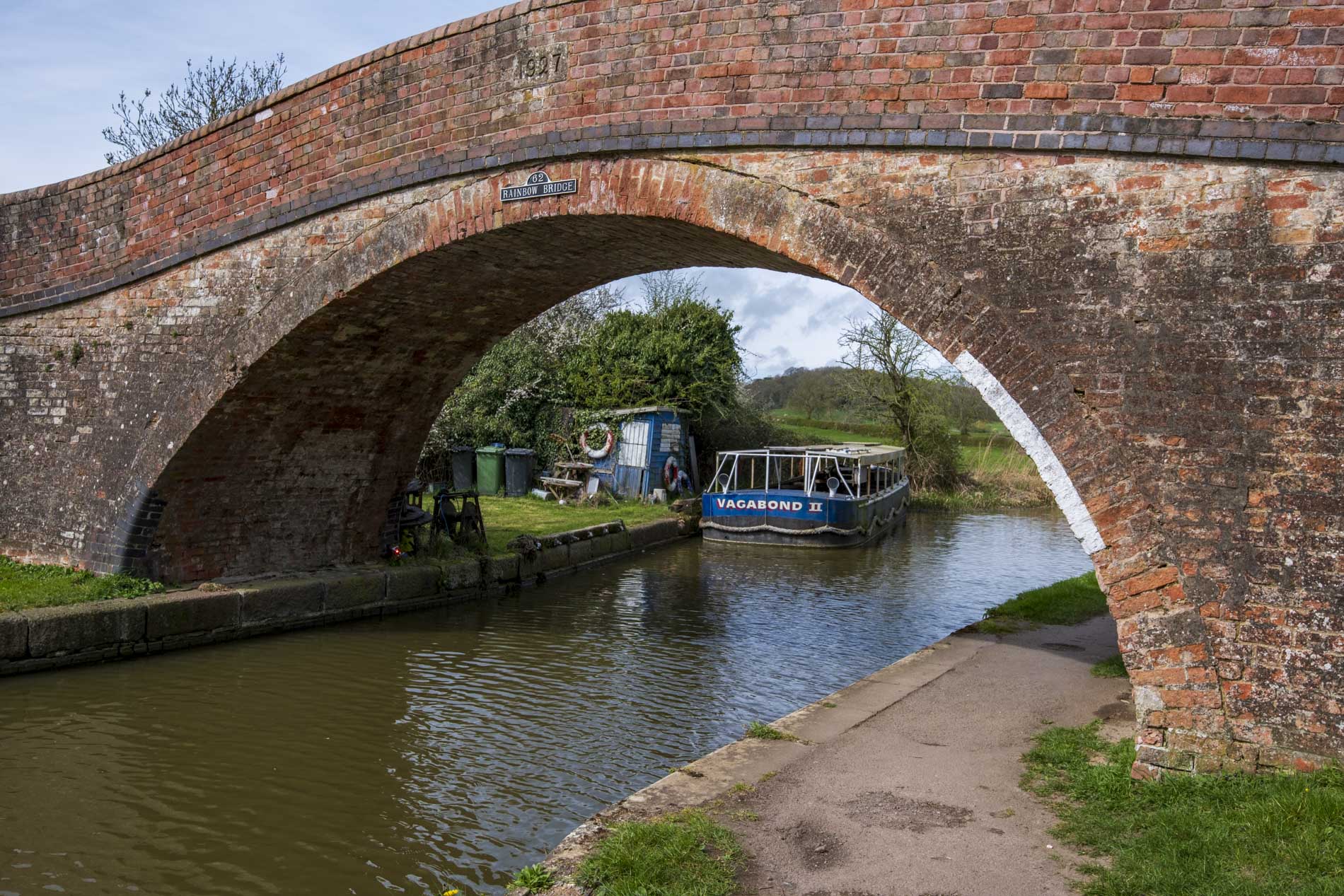 A brick arch bridge spans over a canal, with a small boat named 'Vagabond II' moored nearby. Lush greenery and a shed are visible along the waterway.