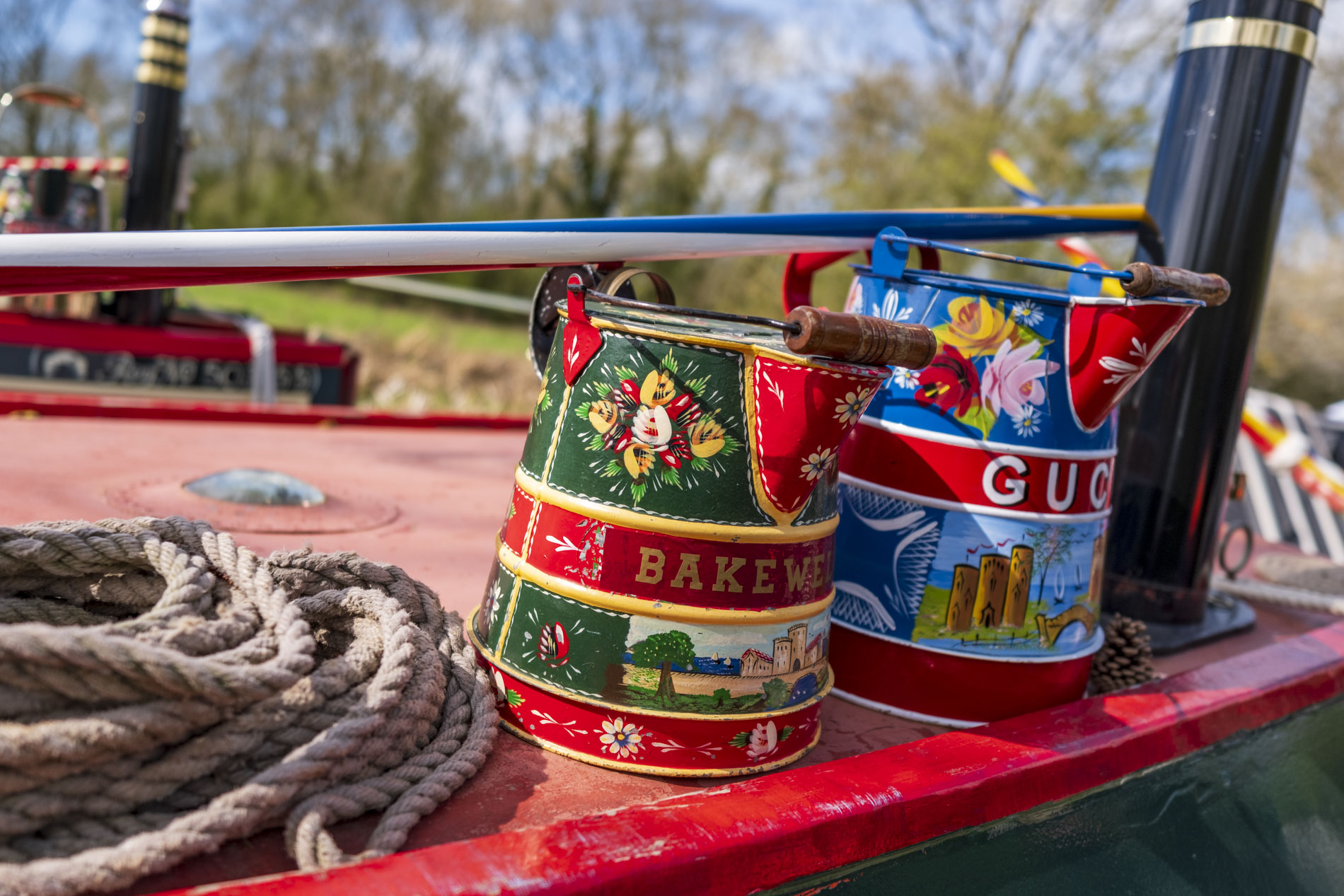 Two decorative watering cans in vibrant colours, one labelled 'BAKEWELL' and the other featuring a floral design and the word 'GUCCI', placed on the edge of a narrowboat deck with a coiled rope nearby.