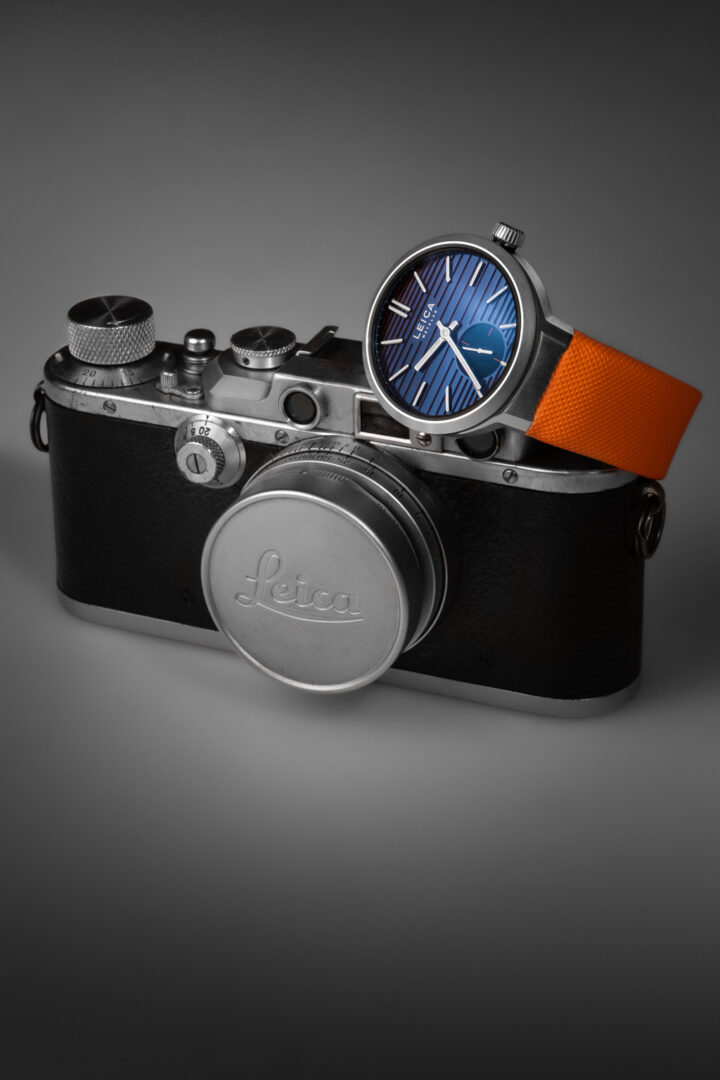 A vintage Leica camera with a classic design, paired with a modern watch featuring a blue dial and an orange strap.