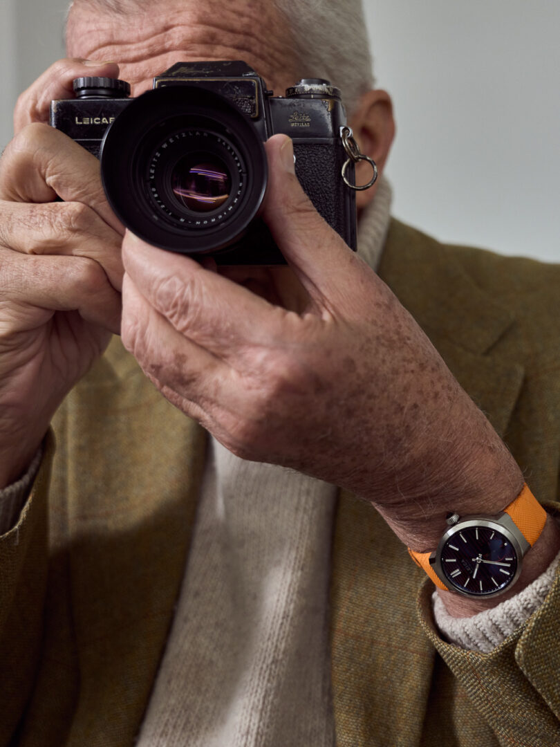 An older man holding a camera in both hands, preparing to take a photo. He is wearing a cream-colored sweater and an olive green blazer, and has an orange watch on his wrist.
