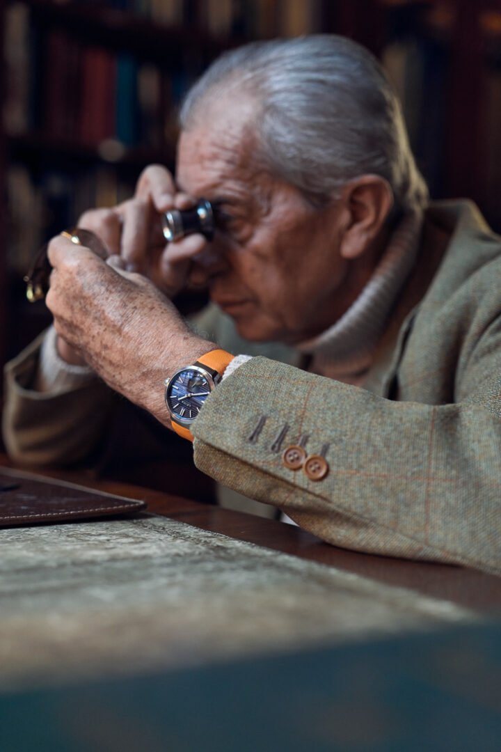 An elderly man examining an object with a magnifying glass while sitting at a table, wearing a stylish watch and a beige blazer.