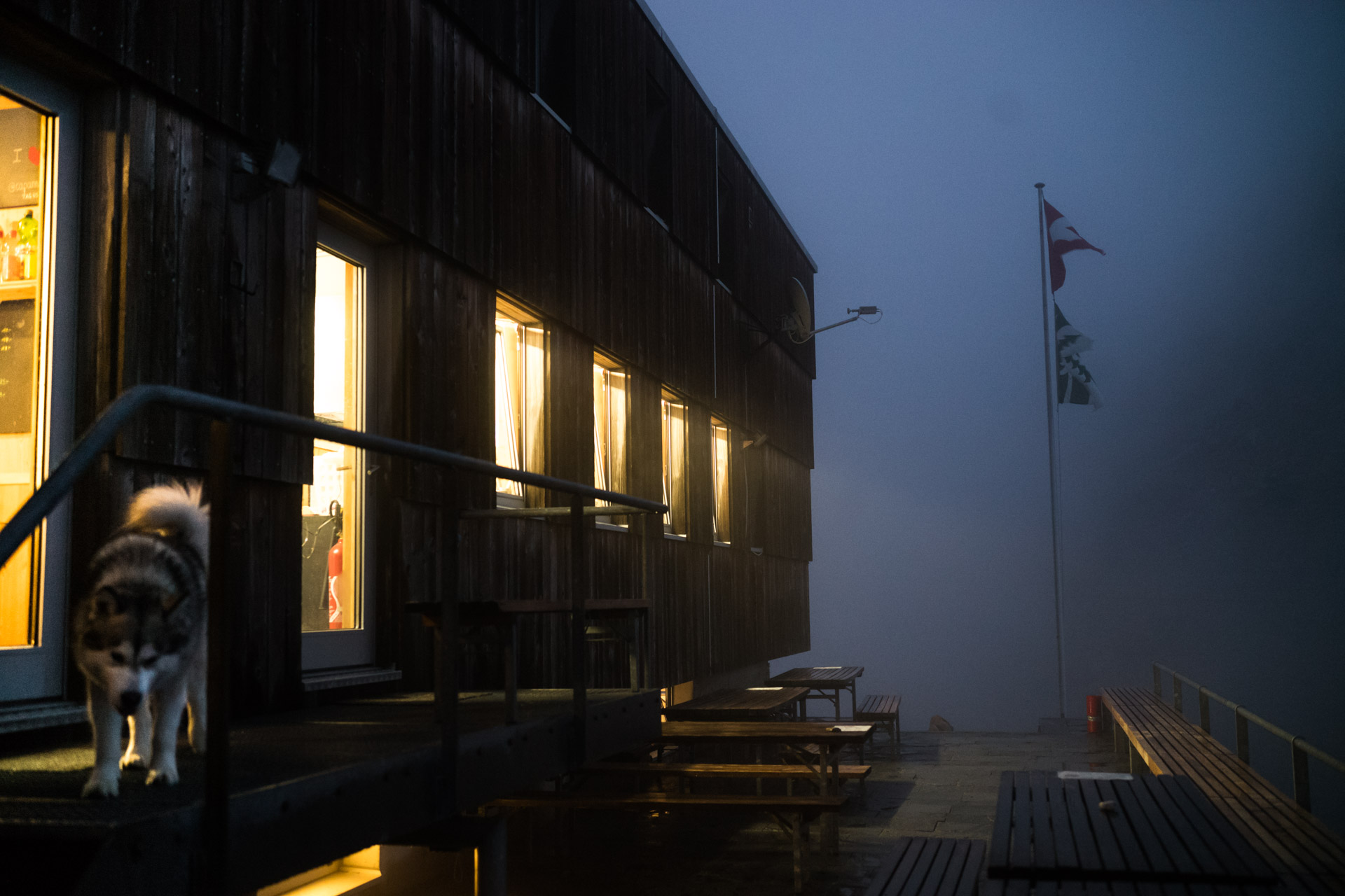A wooden building with lit windows partially shrouded in fog, with a dog walking on the steps. A flag is visible in the background.