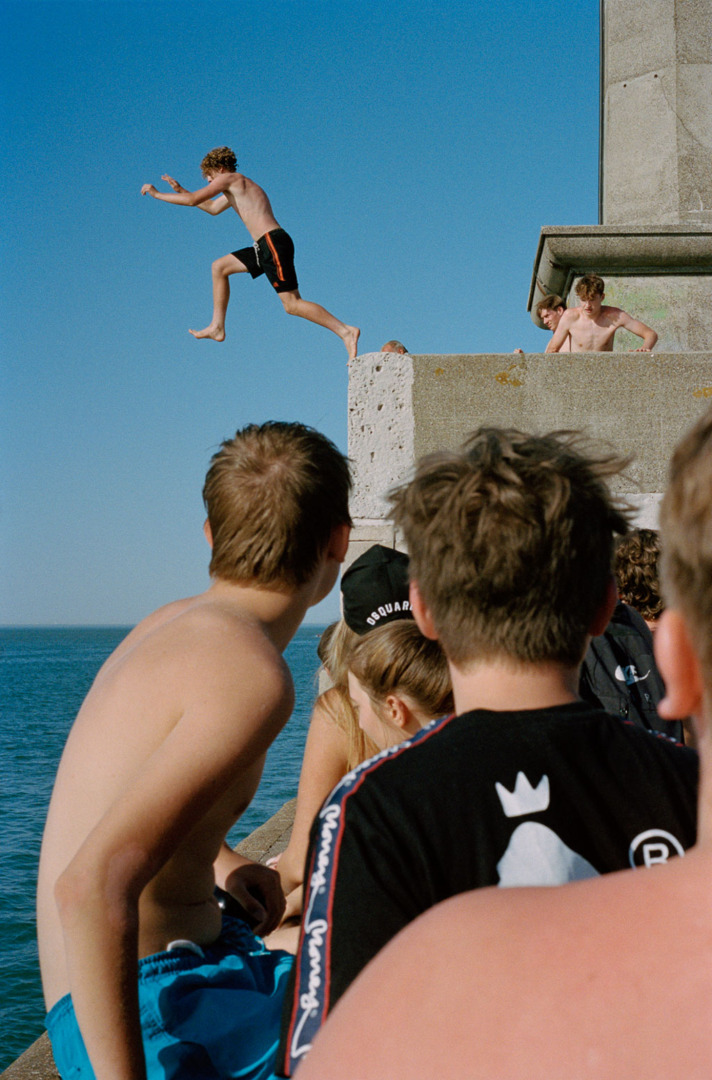 A group of young people watching as a boy jumps off a ledge into the water on a sunny day.