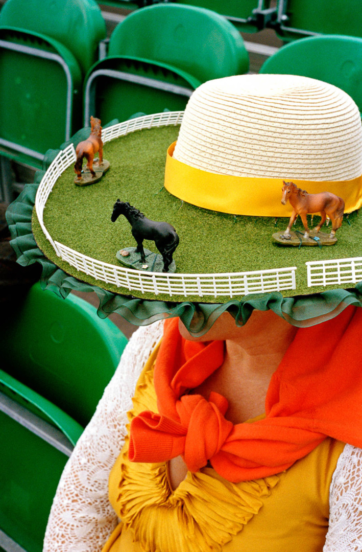 A person wearing a large, decorative hat resembling a horse racetrack, featuring miniature horse figurines and vibrant colors.