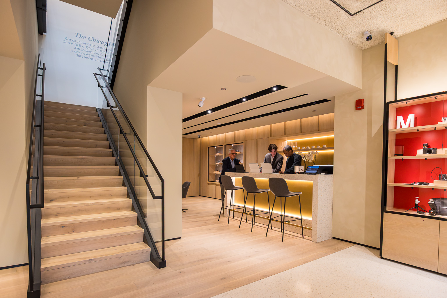 A modern interior of a retail space featuring a wooden staircase leading upstairs, a counter with three people engaged in conversation, and neatly arranged shelves displaying products.