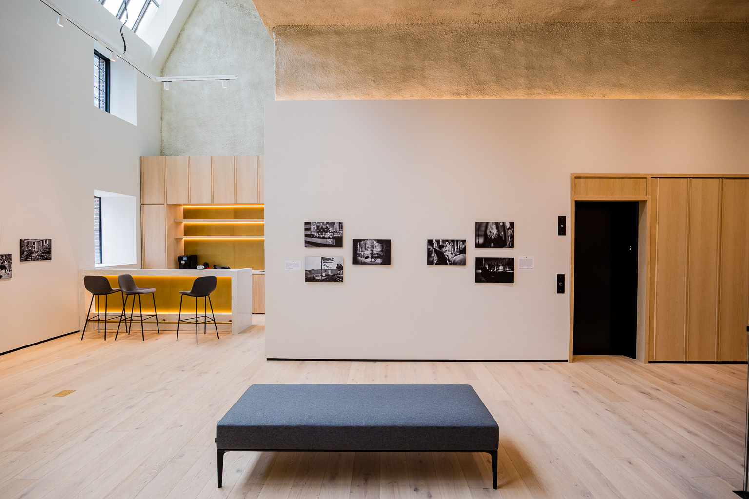 A modern interior space featuring wooden flooring, a minimalist design with a gray sofa in the center, a bar area with stools, and black-and-white photographs displayed on the walls.