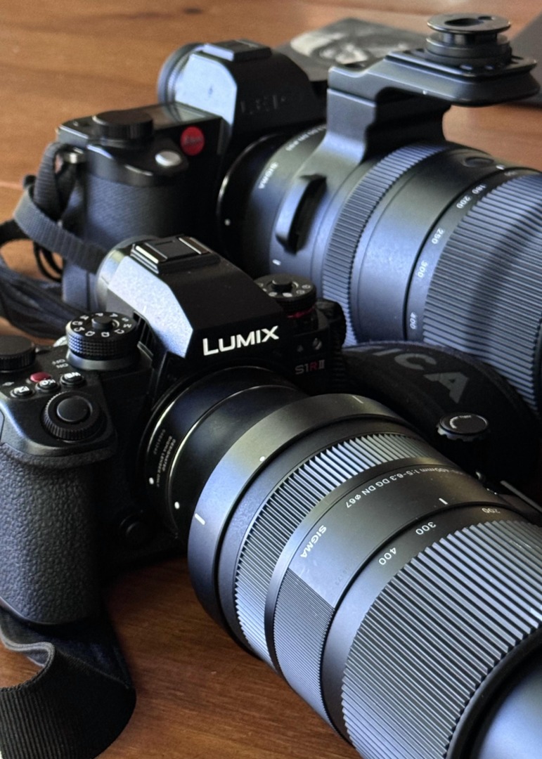 Close-up of three camera bodies on a wooden surface, featuring a Lumix S1R with a Sigma lens, alongside a Leica camera.