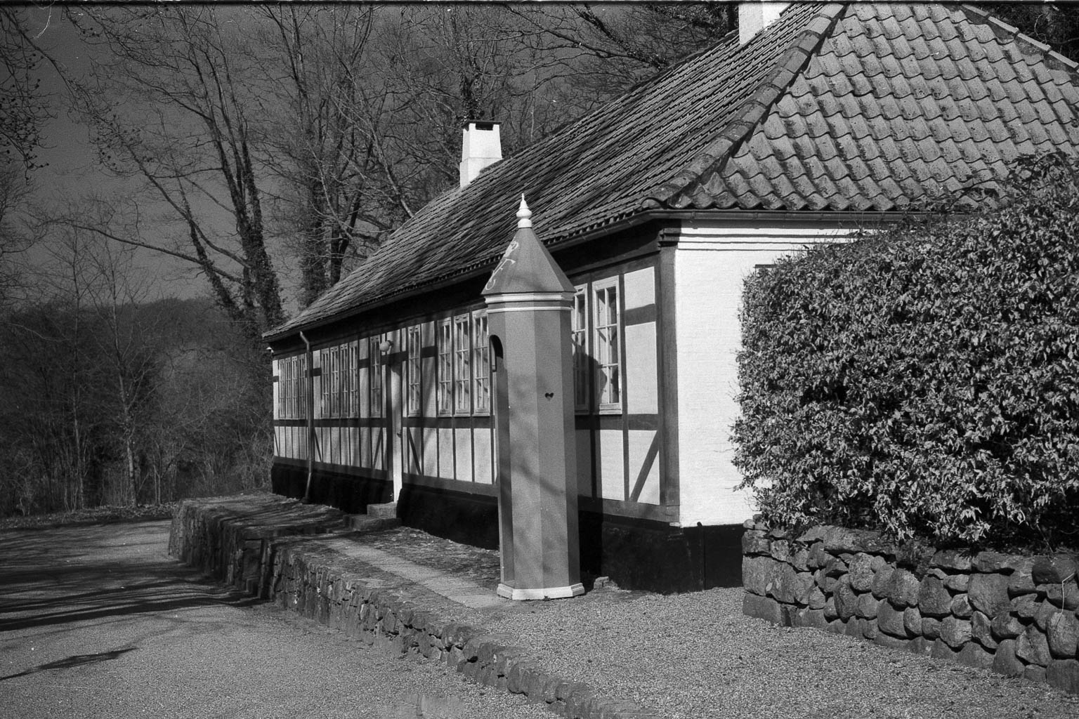 A black and white image of a traditional building with a thatched roof, featuring timber framing. In front of the building, there is a distinctive red kiosk or telephone booth-like structure surrounded by trees and shrubs.