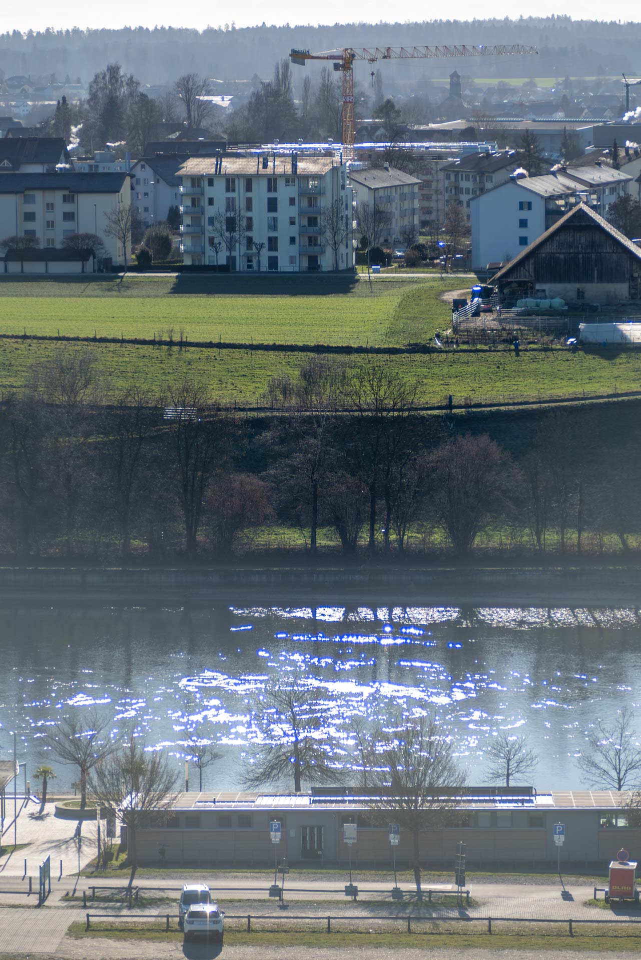 Scenic view of a riverbank with a modern building under construction in the background, surrounded by grassy fields and trees.