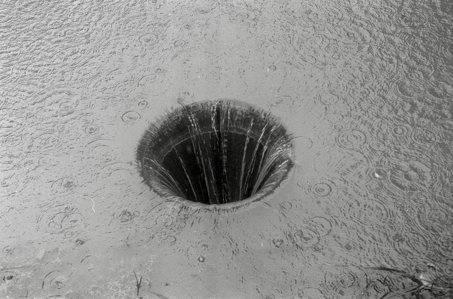 A black and white image depicting a circular drainage hole in a surface, surrounded by ripples from falling rain.