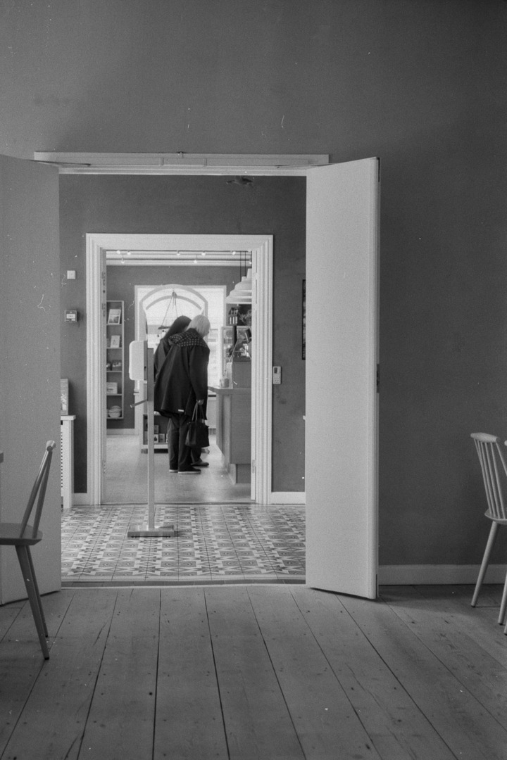 A view through an open doorway into a room featuring tiled flooring, with two figures engaged in conversation near a table in the background. The image is in black and white.